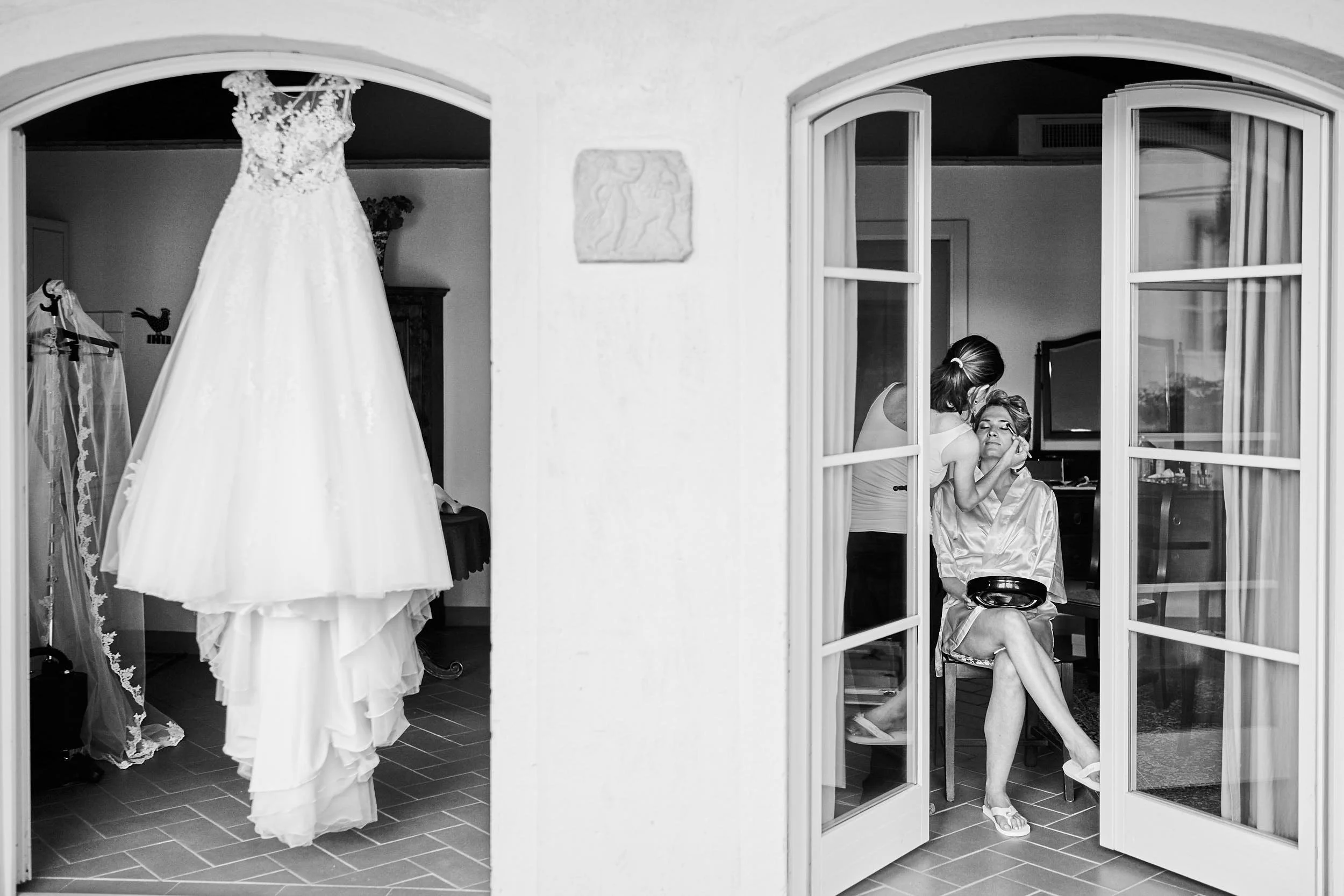 Black-and-white composition showing the wedding dress hanging on the left and the bride getting makeup on the right, seen through open doors.