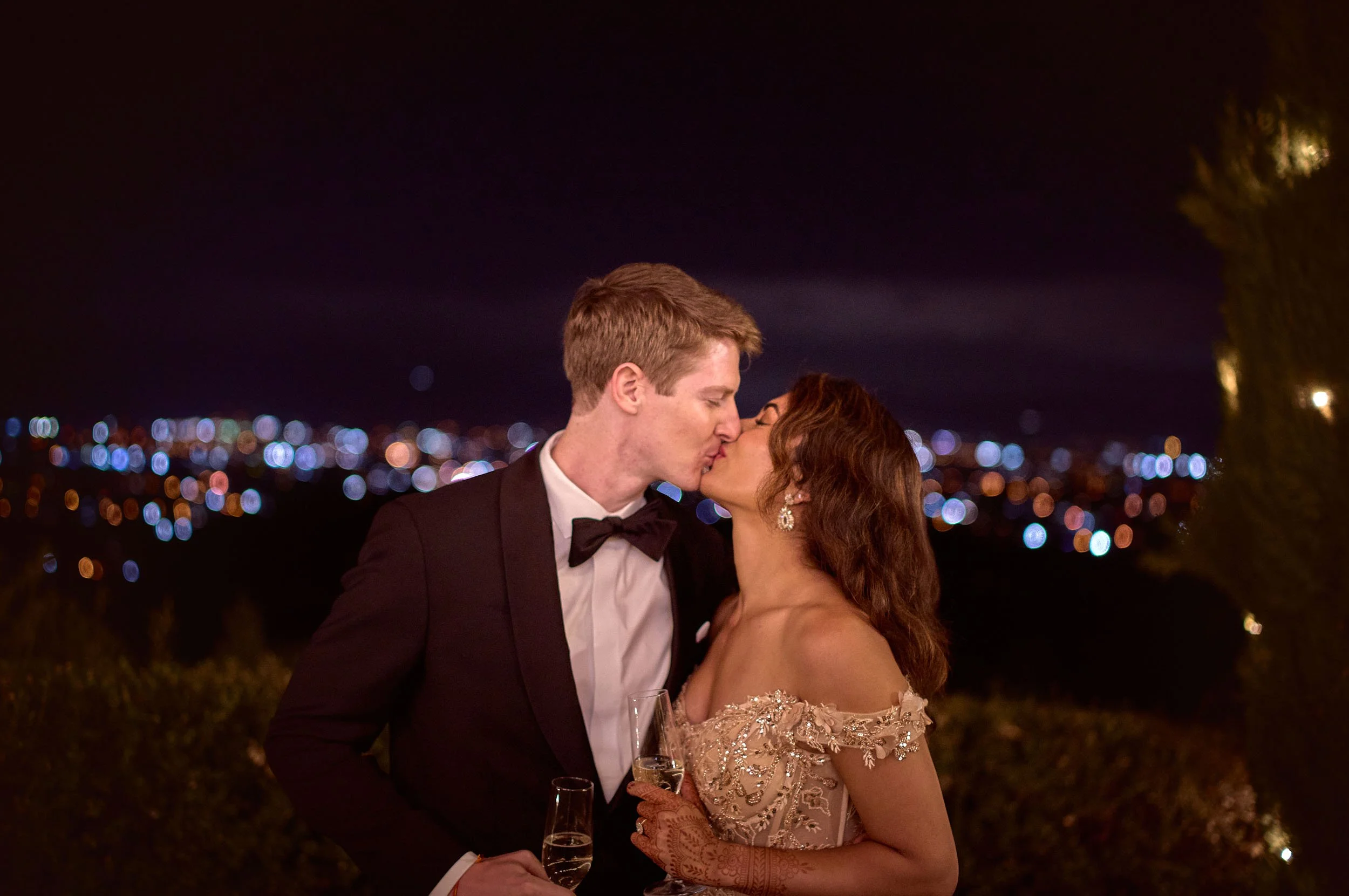 Bride and groom kiss after cutting the cake — blurred city lights of Lucca behind them