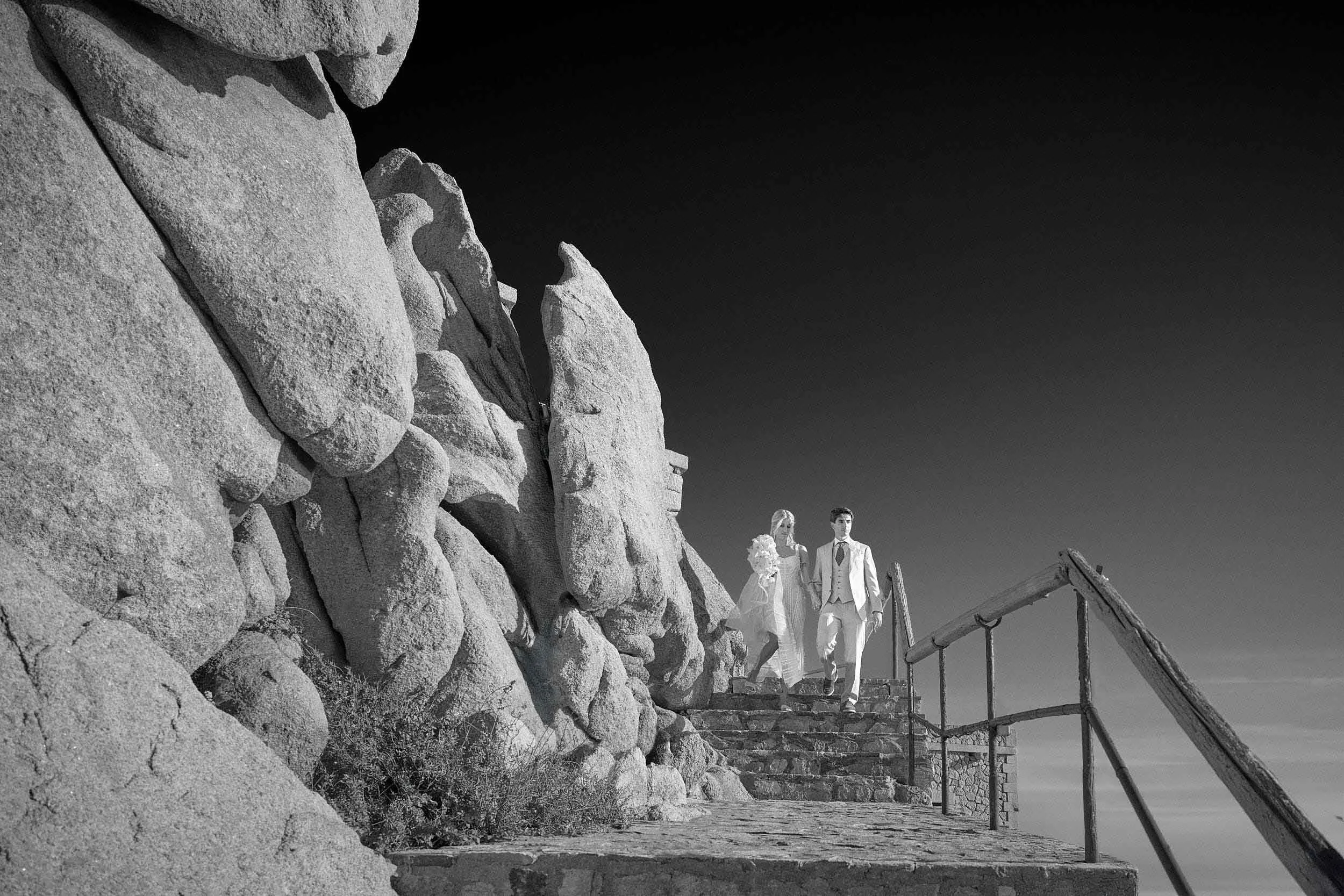 A bride and groom walking down the stone stairs at The Rock, Phi Beach private club, surrounded by massive granite rock formations under a clear sky. High-contrast black and white wedding photography in Sardinia.