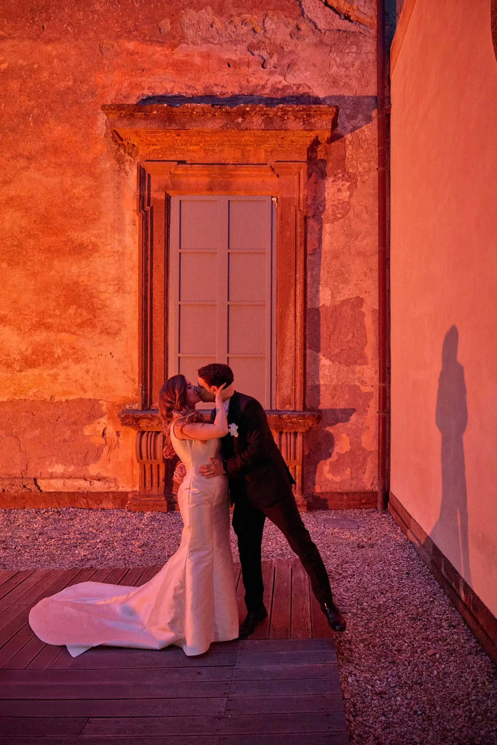 Bride and groom kiss at night in front of a window at Villa Mondragone; their shadow is cast to the right by a villa light.”