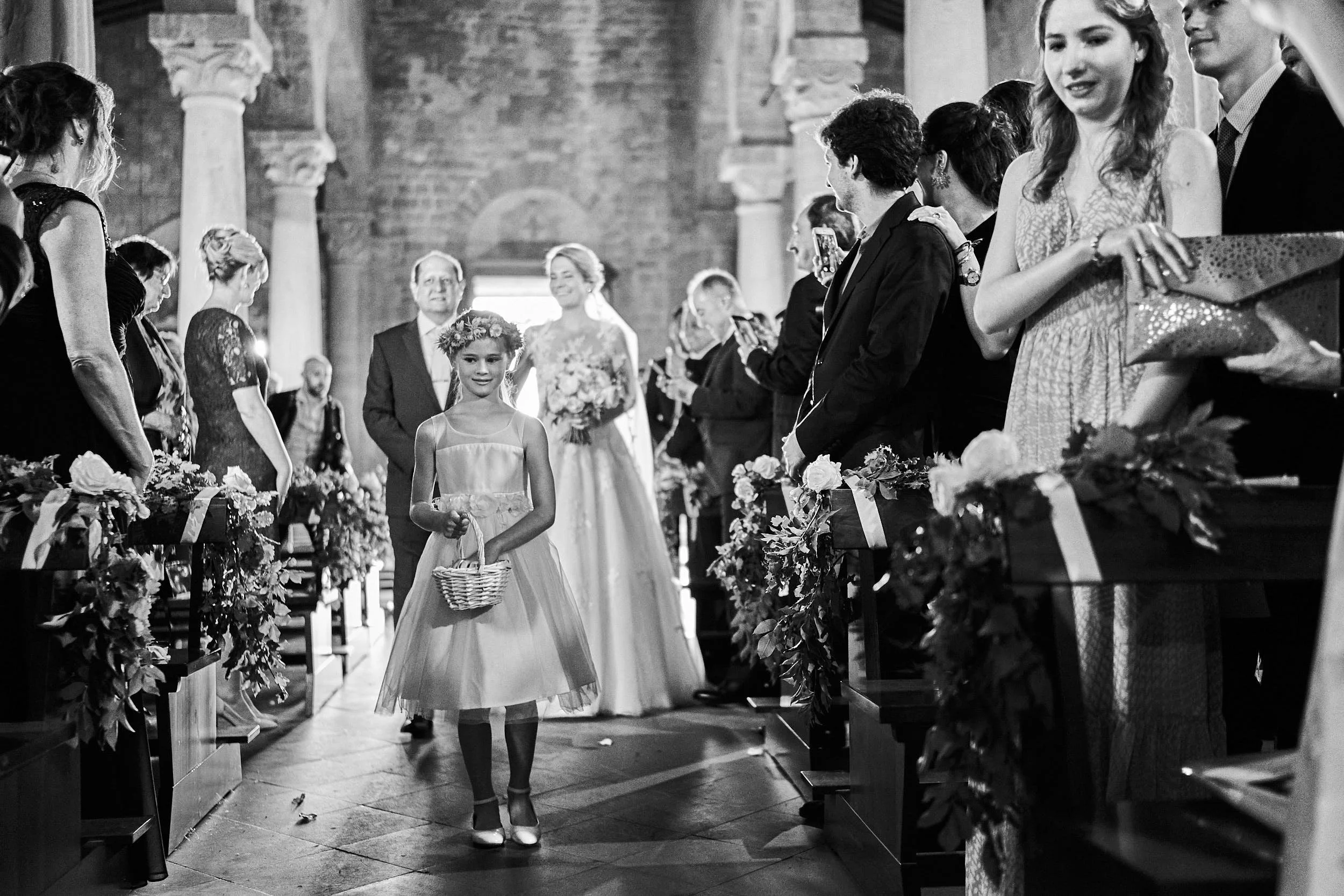 Flower girl walks down the church aisle as guests watch, with the bride arriving in the background (black and white).