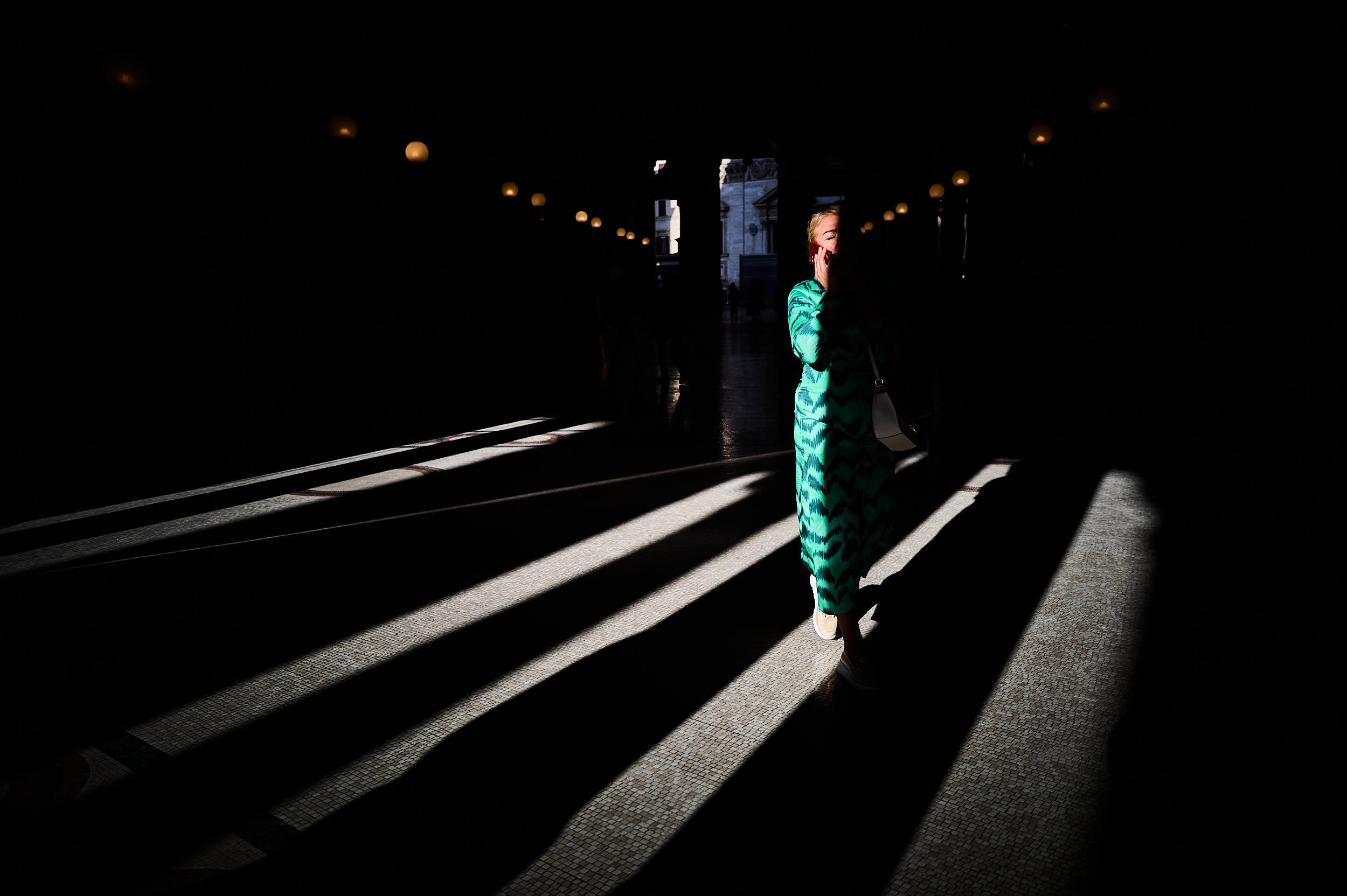 A woman wearing a green dressing walking between deep shadows and harsh light while making a call with her phone