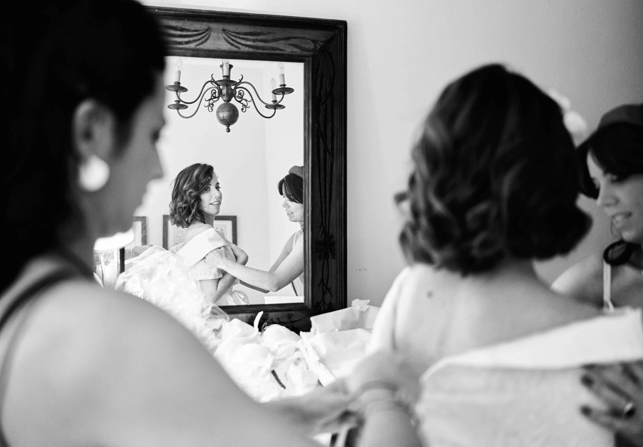 Bride reflected in a mirror while an attendant adjusts her dress during preparations at Tenuta di Polline, Italy.