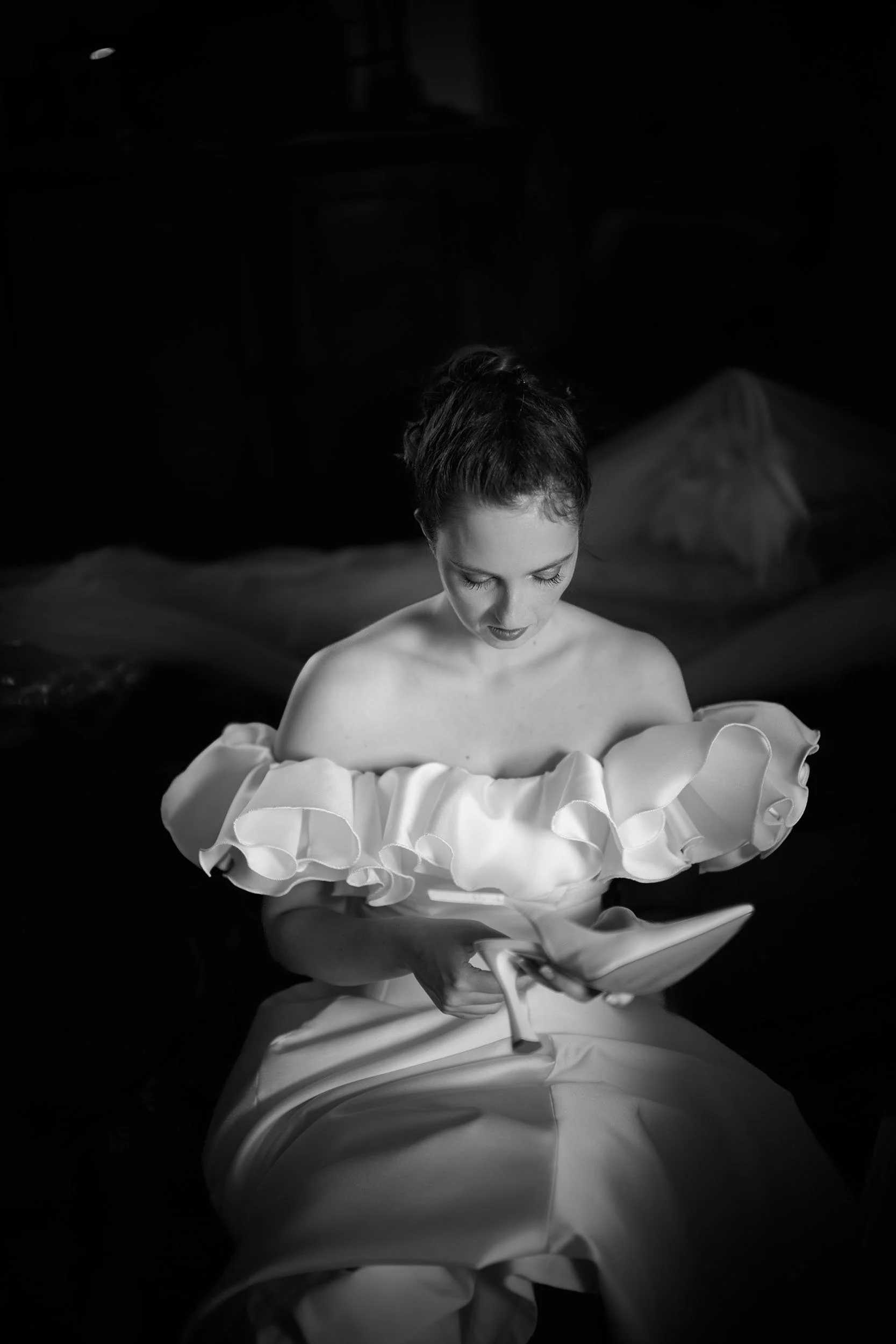 Bride sitting in the shadows looking at her elegant shoe during preparations, an intimate black and white portrait highlighting an unposed documentary and editorial aesthetic.