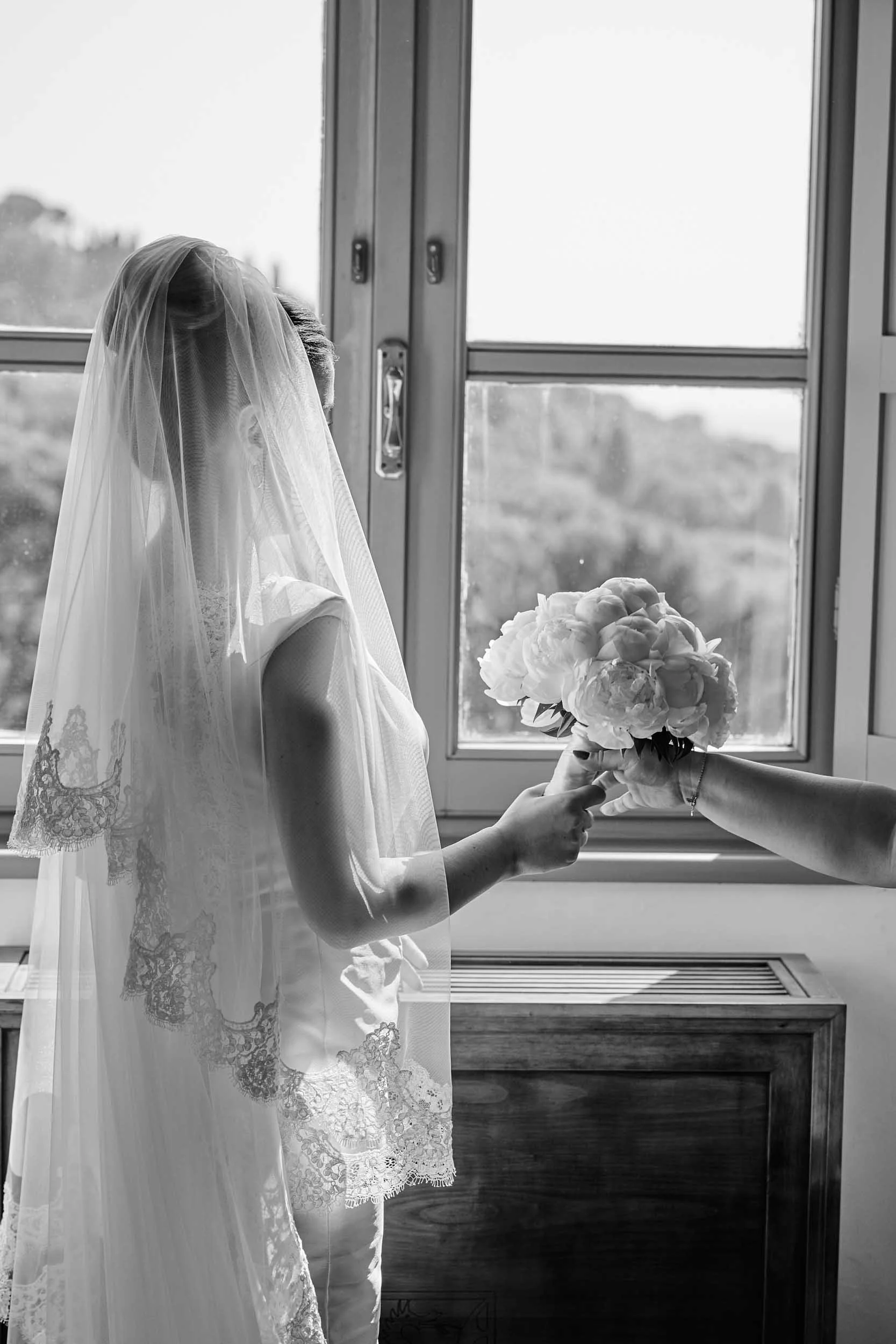 Black-and-white photo of the bride picking up her flower bouquet at the window.