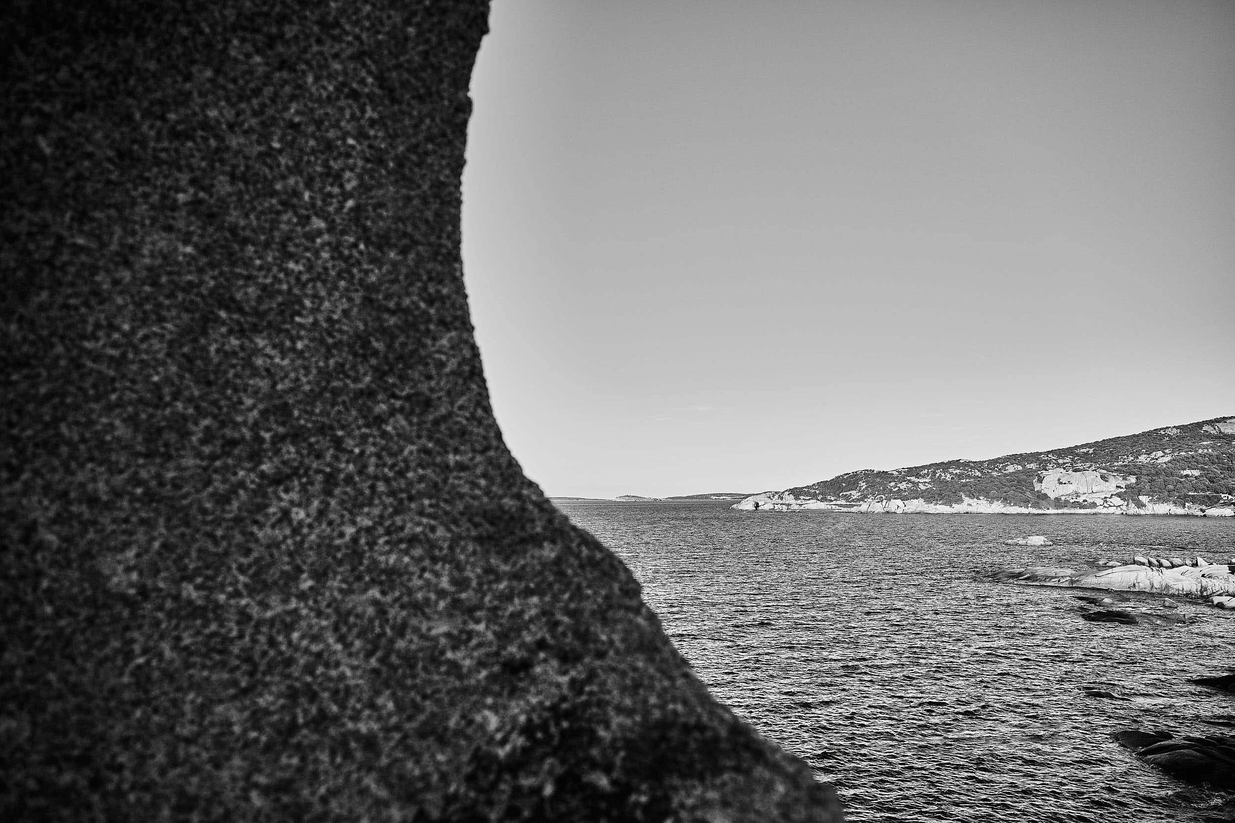 Granite rock in the foreground framing the Mediterranean coastline in black and white, Costa Smeralda, Sardinia.