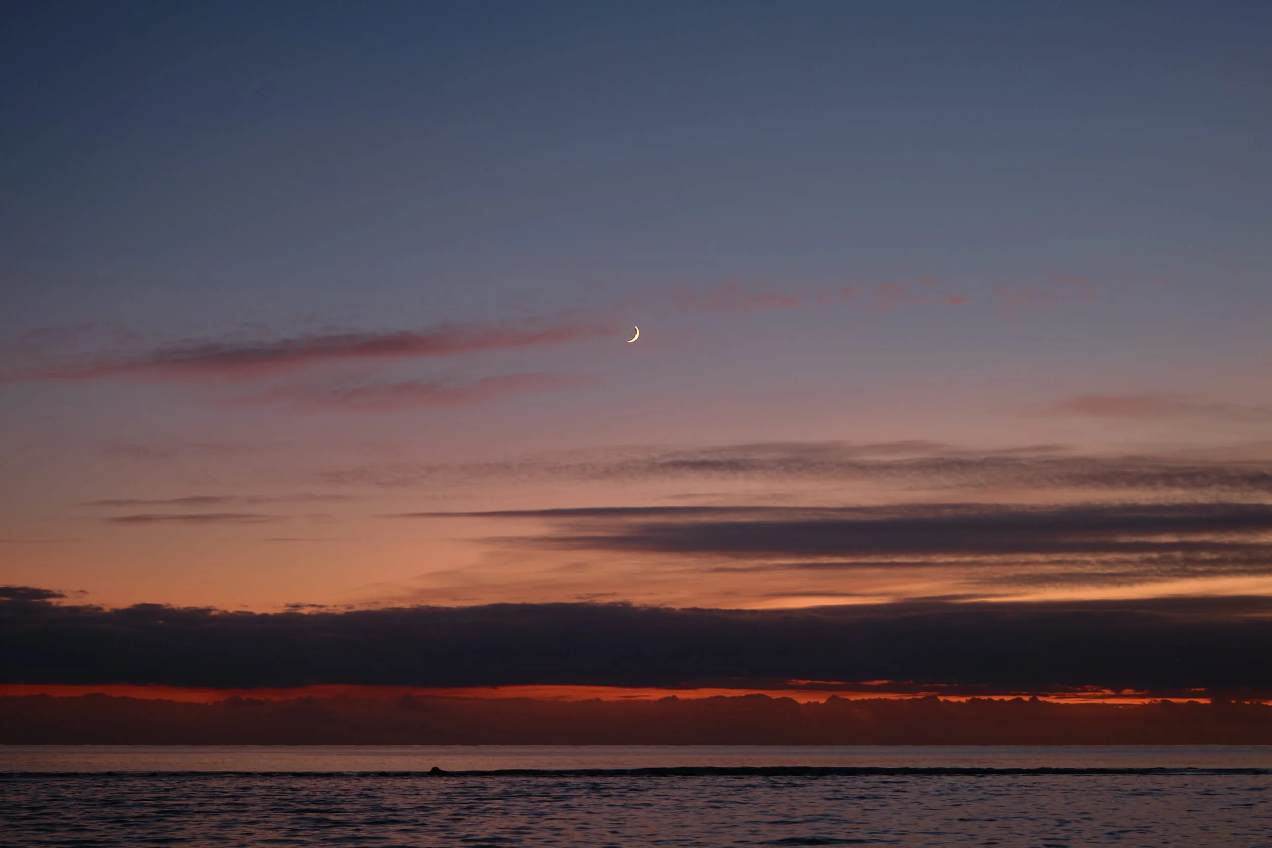 Minimal seascape at dusk with a thin crescent moon above layered clouds and a deep orange glow along the horizon.