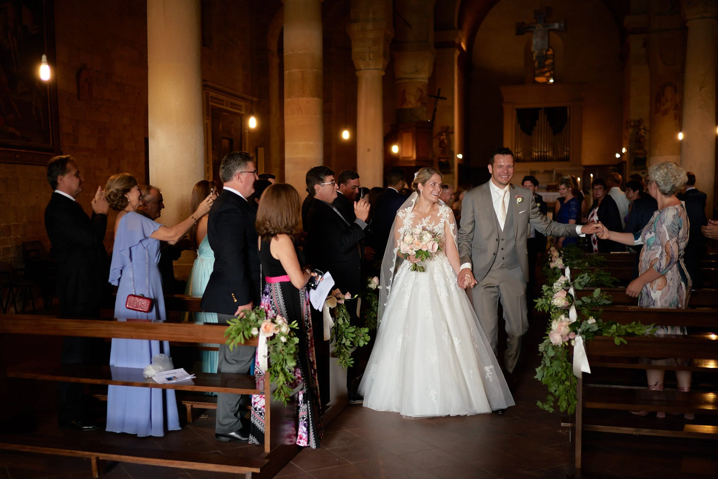 Bride and groom walk down the aisle inside a church as guests applaud, a documentary moment with warm tones and natural reactions.