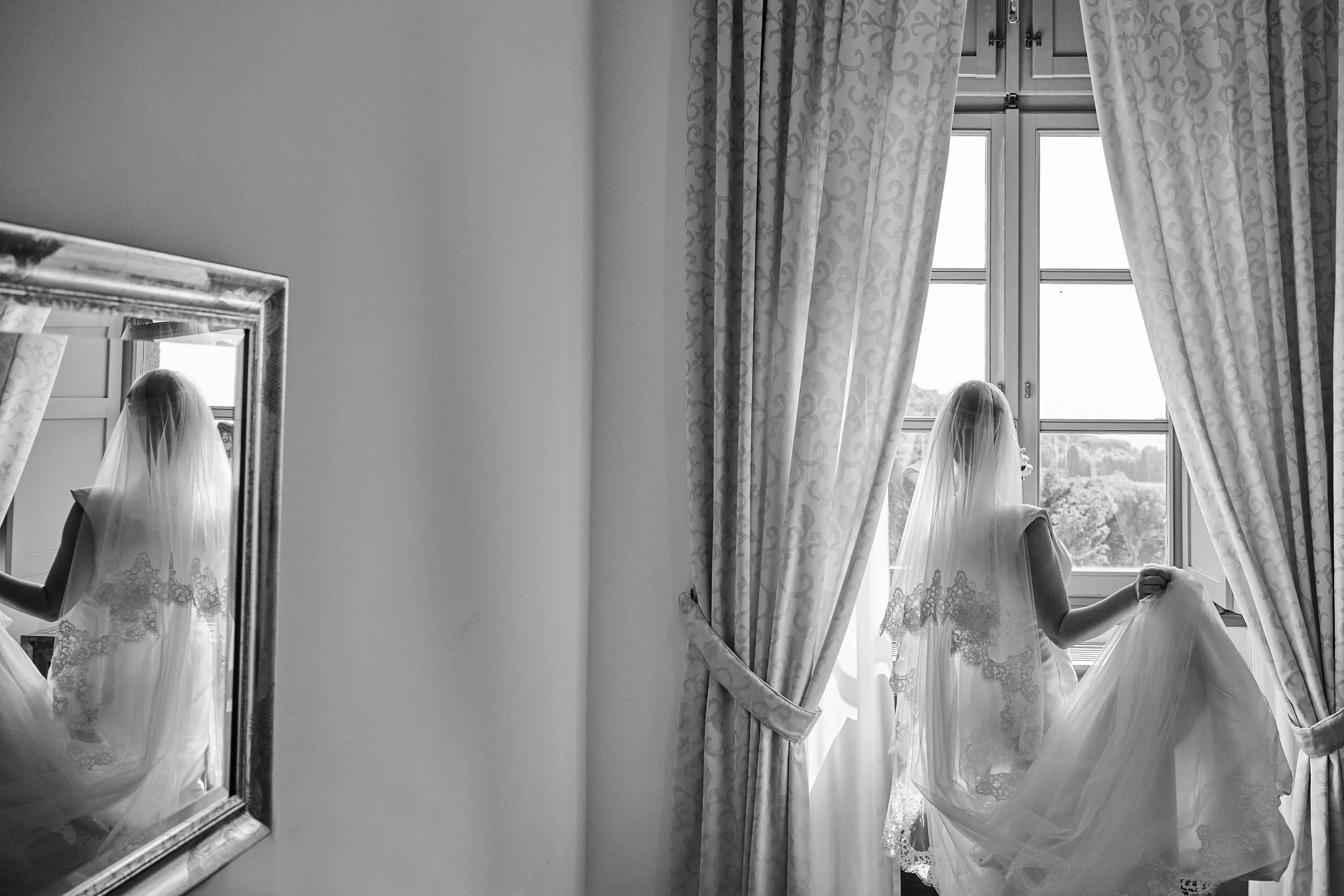 Black-and-white: bride gazes out the window, reflected in a mirror to her left.