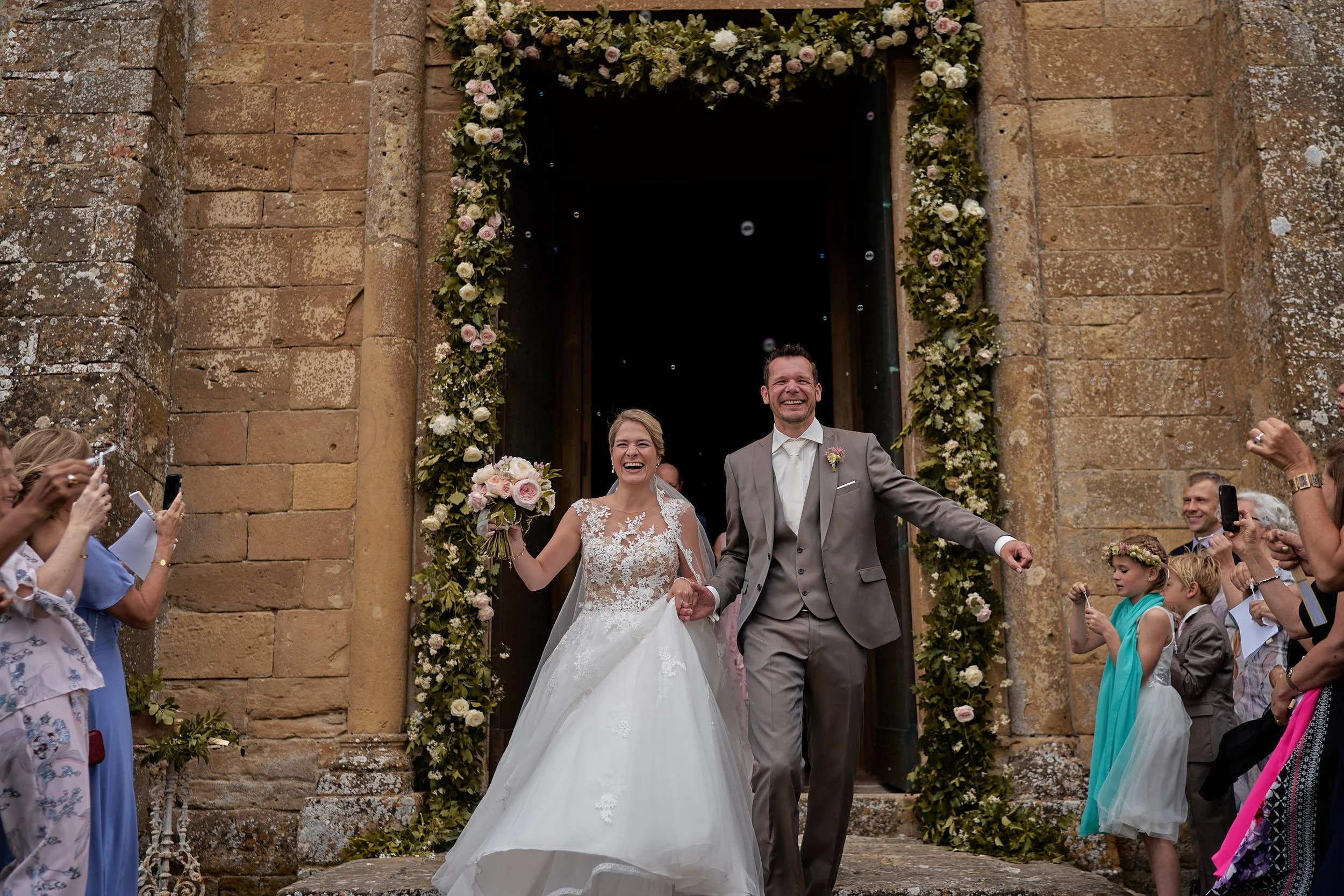 The couple exits the church smiling and holding hands, surrounded by guests and floral details—classic destination wedding energy in Italy.