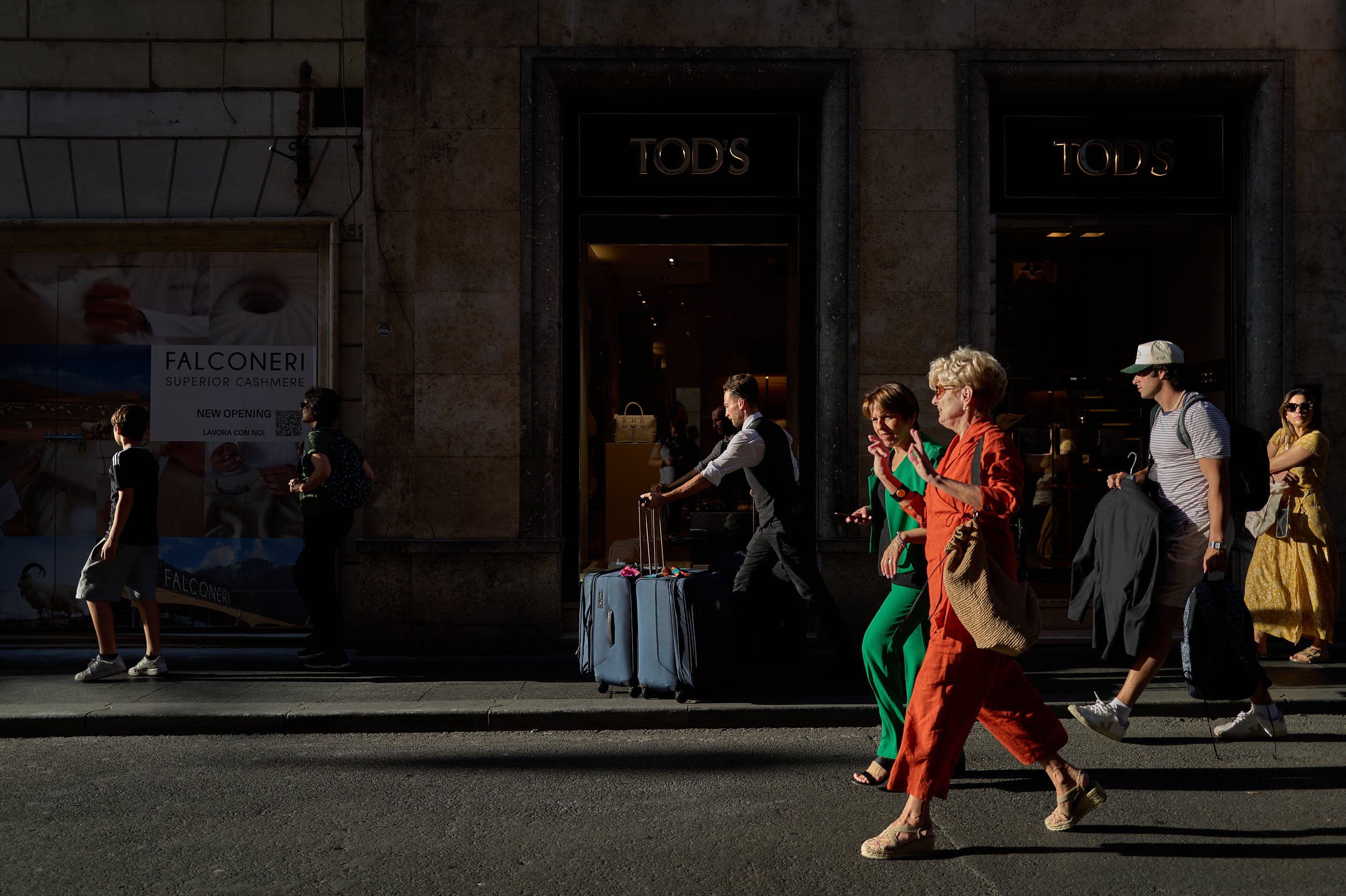 two elegant women walking at sunset in a street in Rome, a man walking with heavy bags in the background