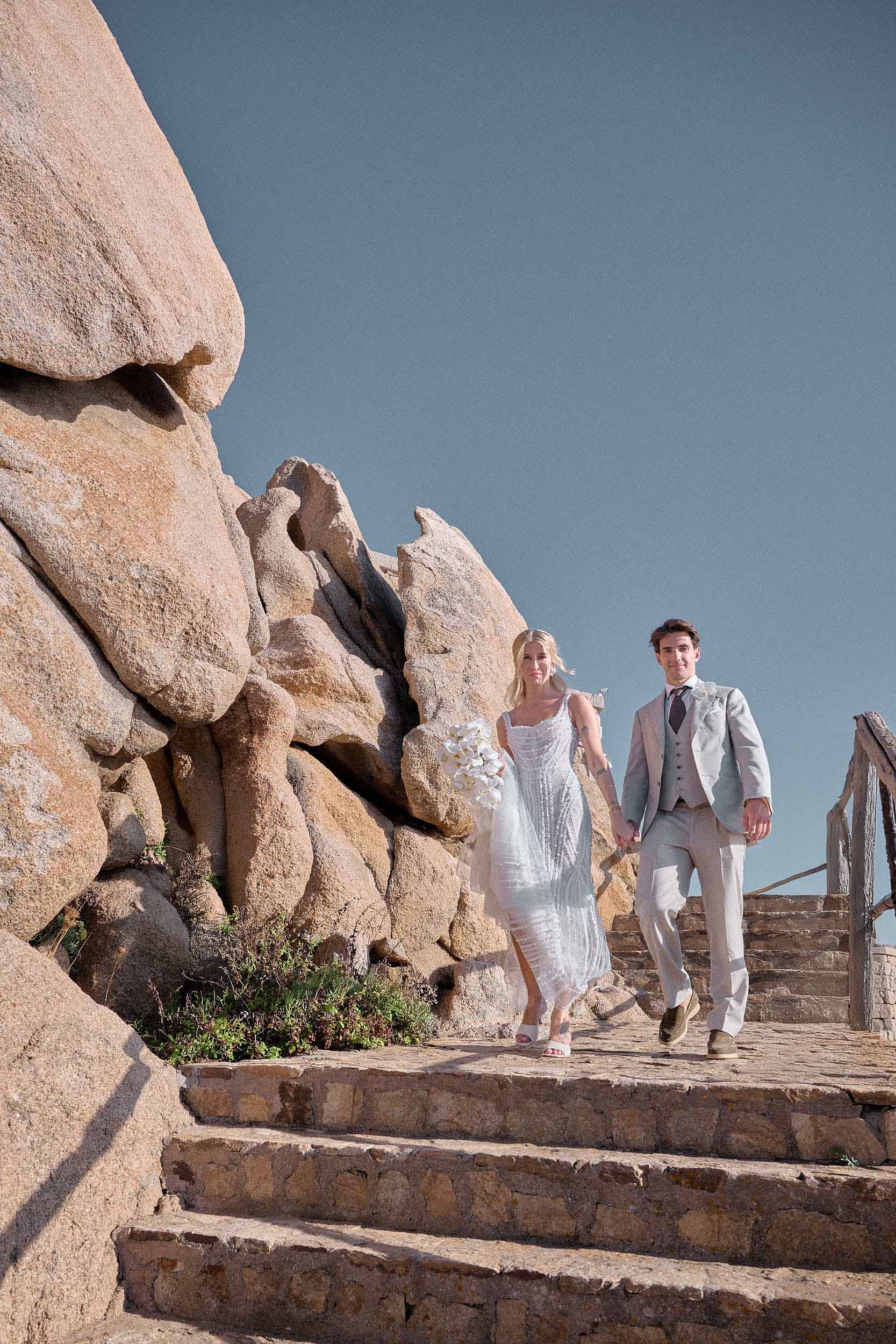 Bride and groom walking down stone steps among granite rocks at Phi Beach “The Rock” in Porto Cervo, Costa Smeralda.