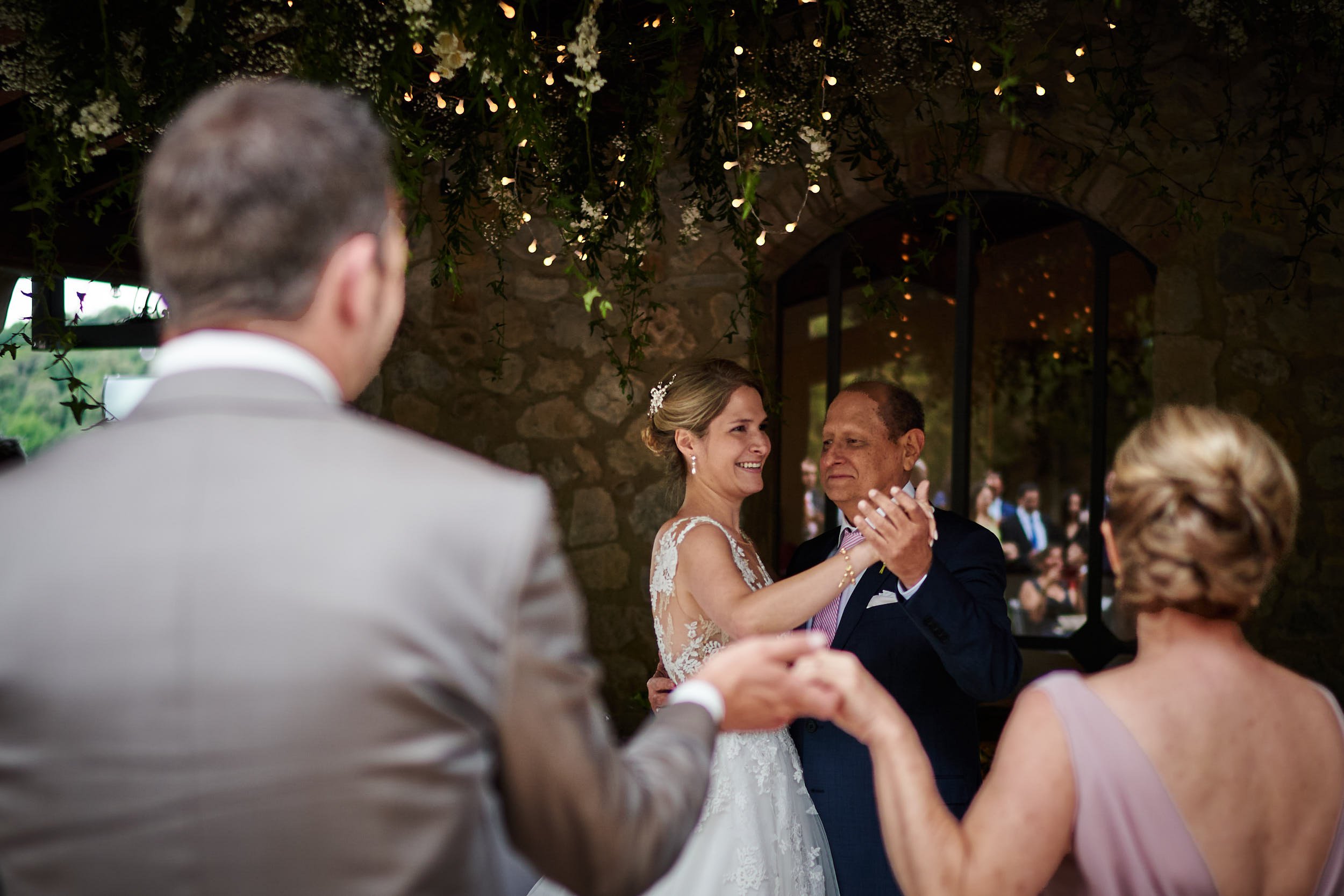 Bride shares a dance with her father beneath hanging greenery and warm string lights, framed by guests in the foreground for depth.