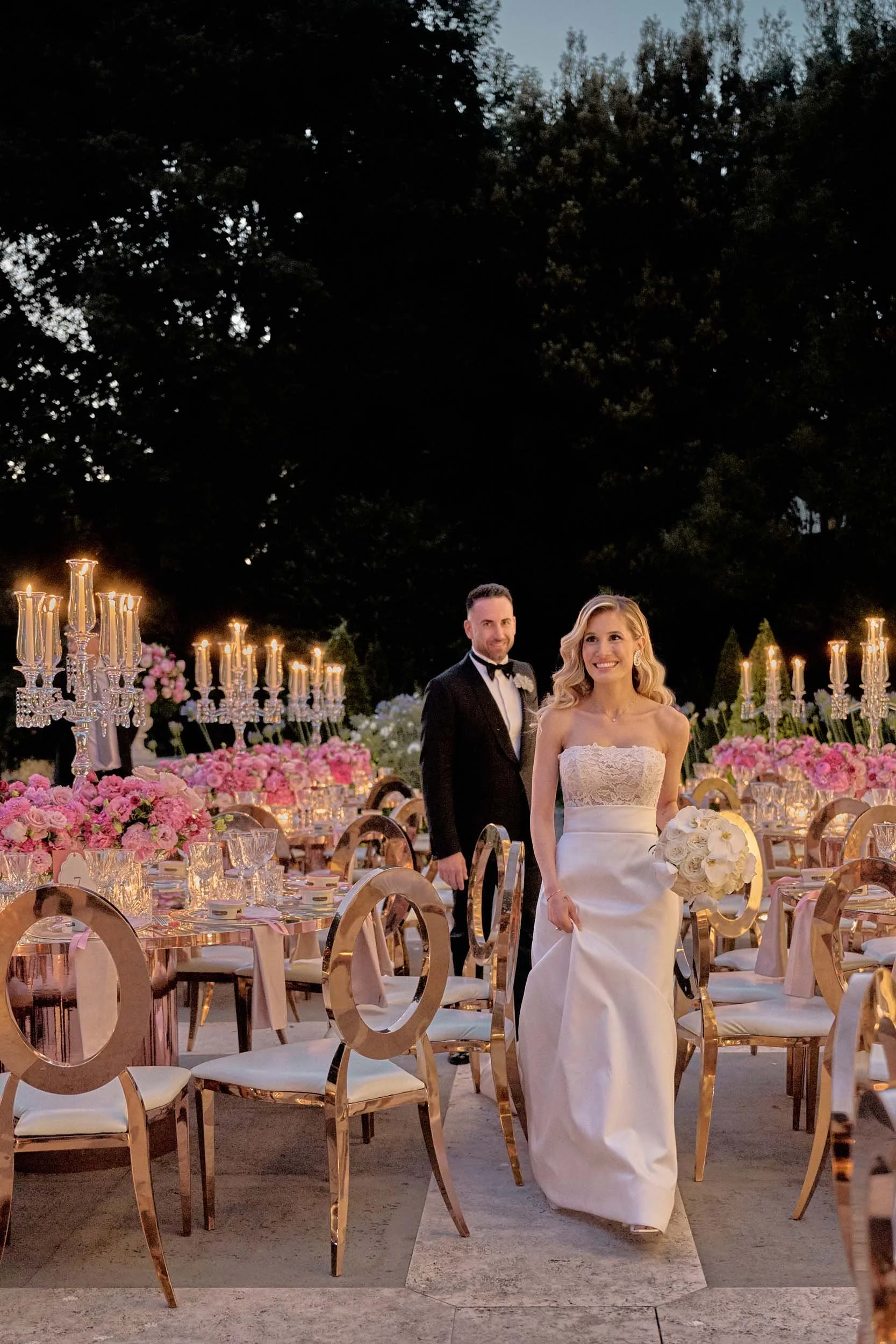 Smiling bride and groom walking past a luxury outdoor floral setup at dusk, elegant photojournalistic  editorial style for an exclusive destination wedding in Italy.
