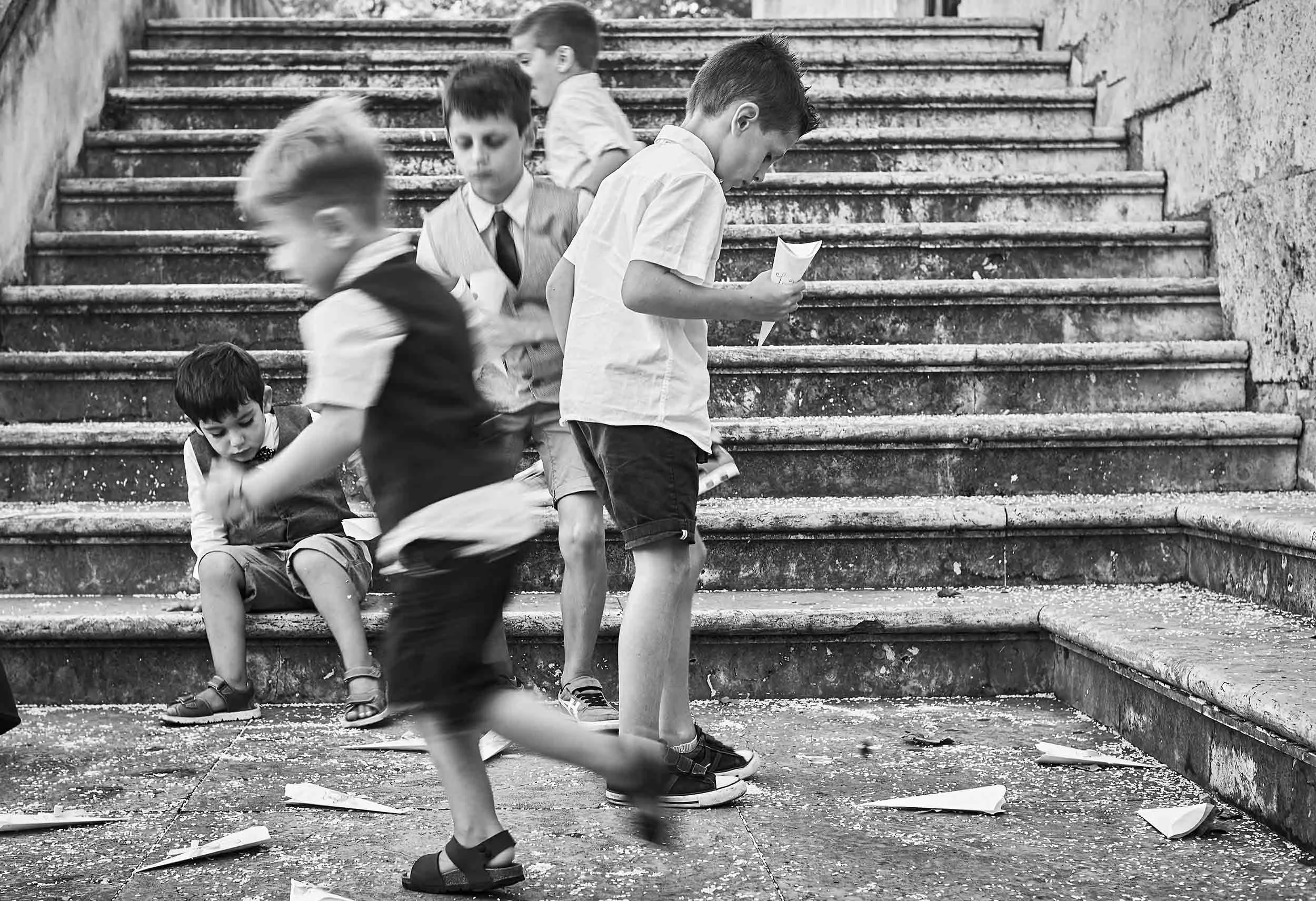 Black-and-white scene of children running and folding paper planes on stone steps during a wedding.