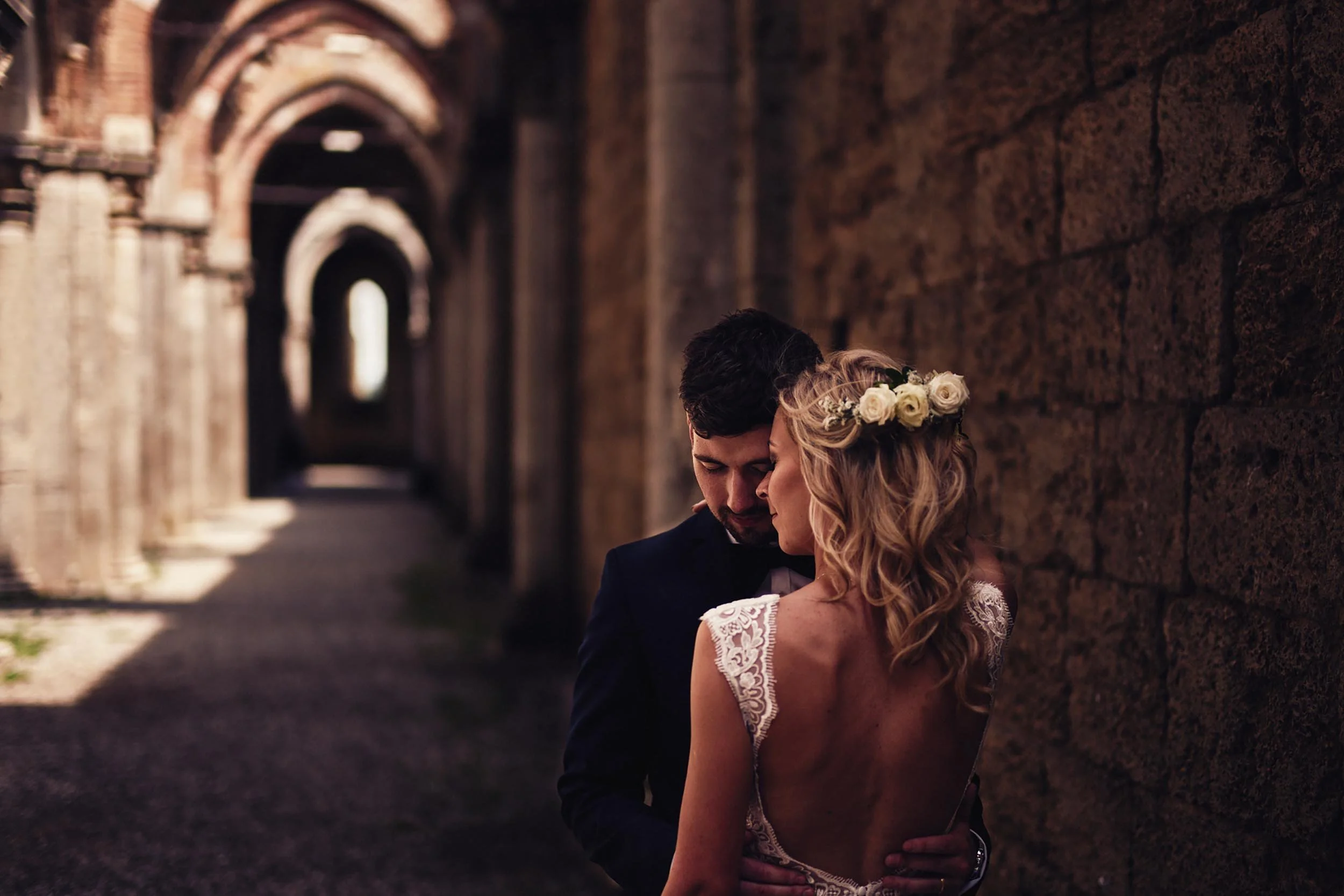 Bride and groom holding each other closely inside an arch of San Galgano Abbey, eyes closed with emotion