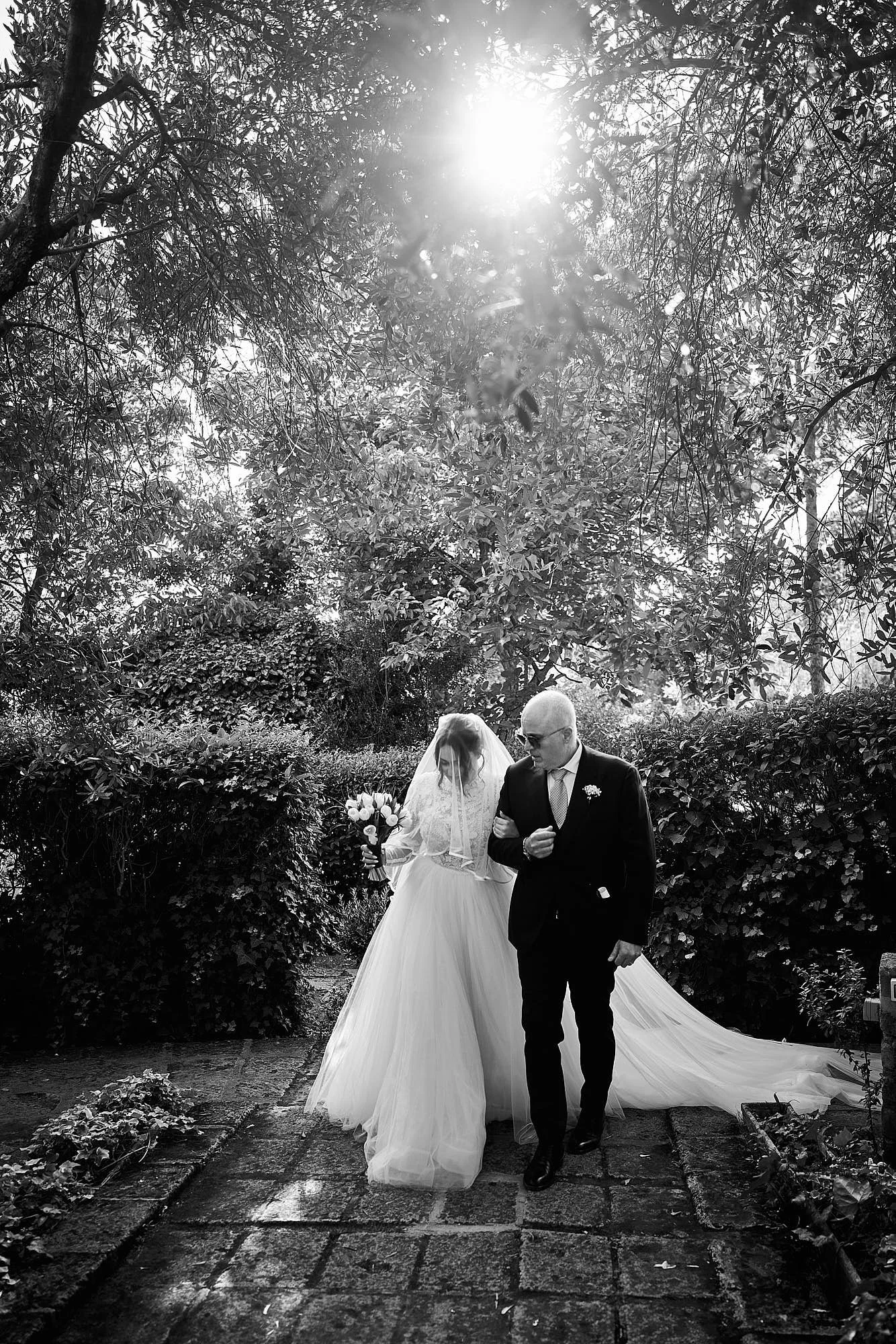 Black-and-white portrait of the bride walking arm-in-arm with her father along a garden path, backlit by the sun filtering through branches.
