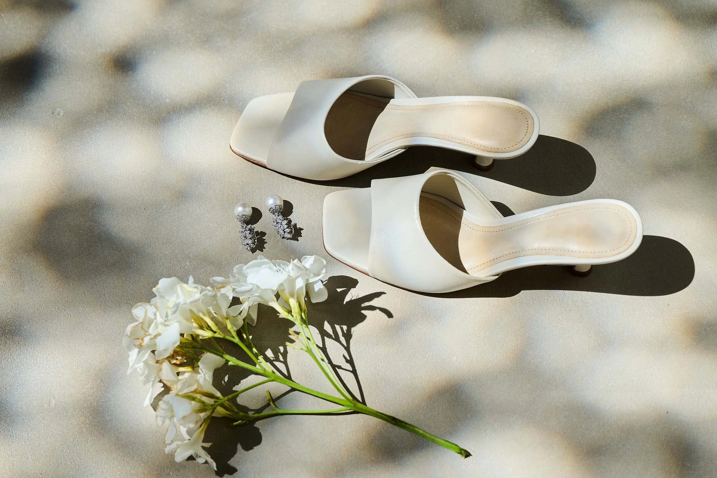 Bridal details—white heeled sandals, pearl-and-diamond earrings and white flowers on a sunlit stone floor, wedding day.