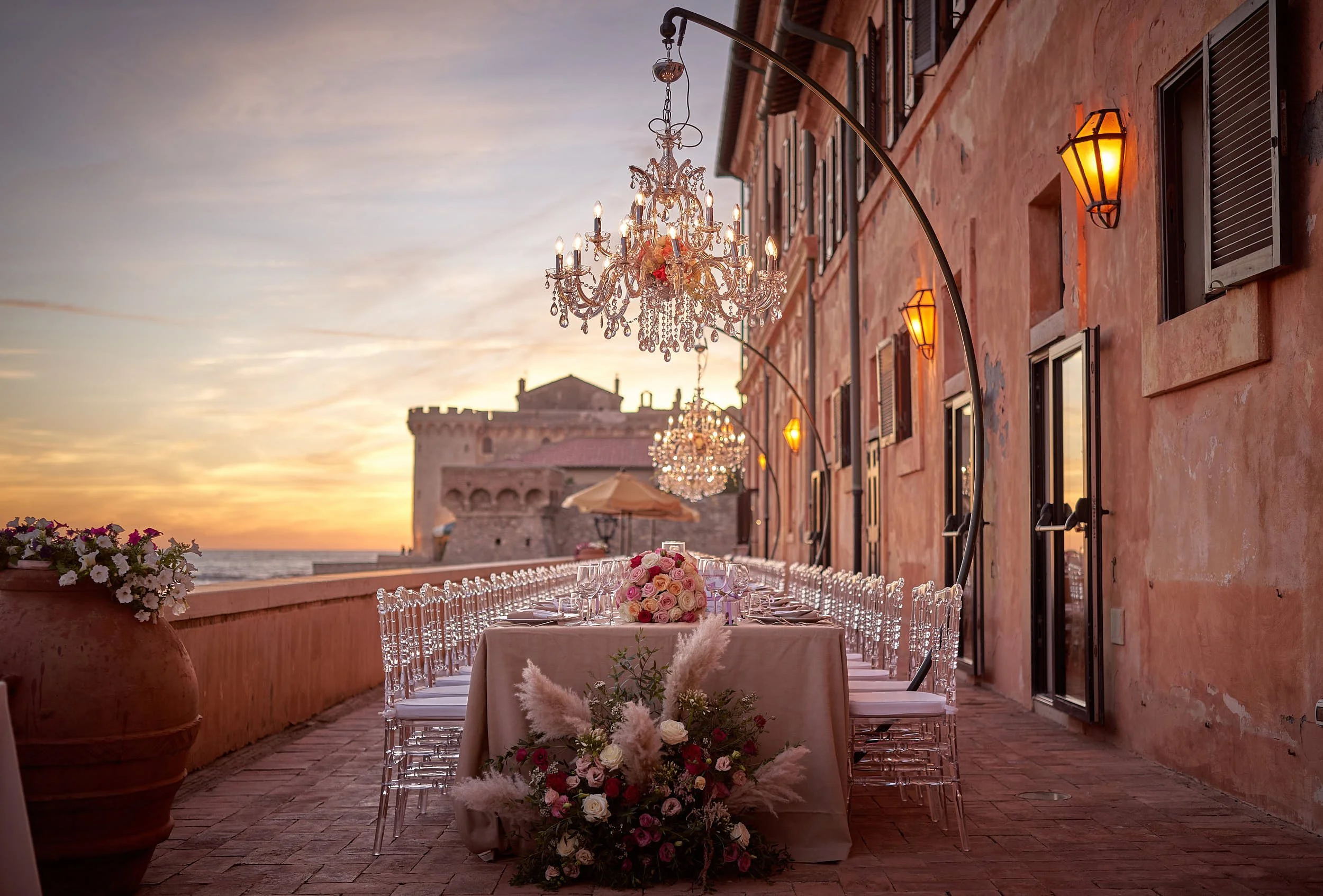 Long banquet table set on a seaside terrace at sunset, crystal chandeliers and warm lights along the building.