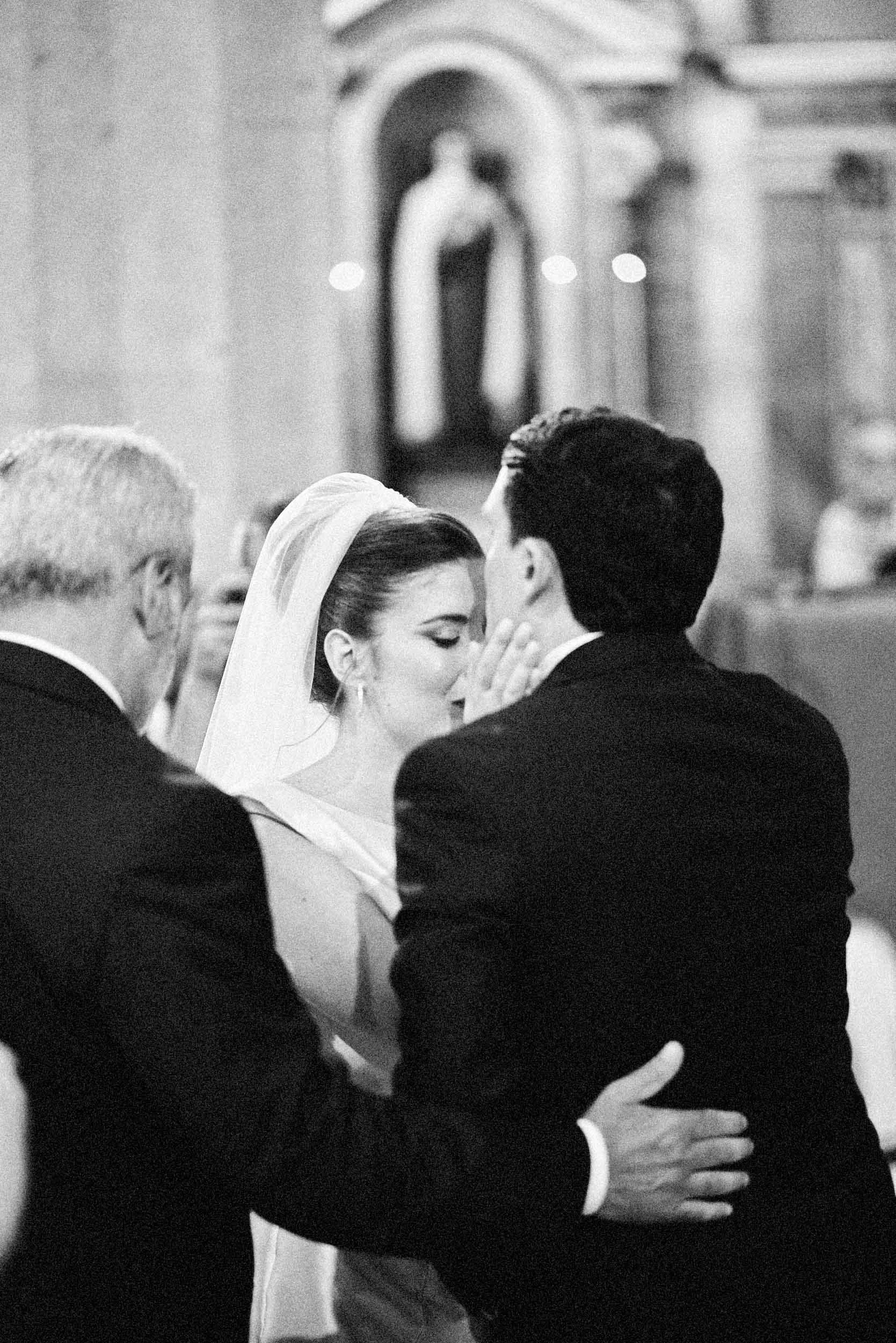 Black-and-white, grainy motion blur: groom kisses the bride’s forehead; she closes her eyes as her father rests an affectionate arm on the groom.