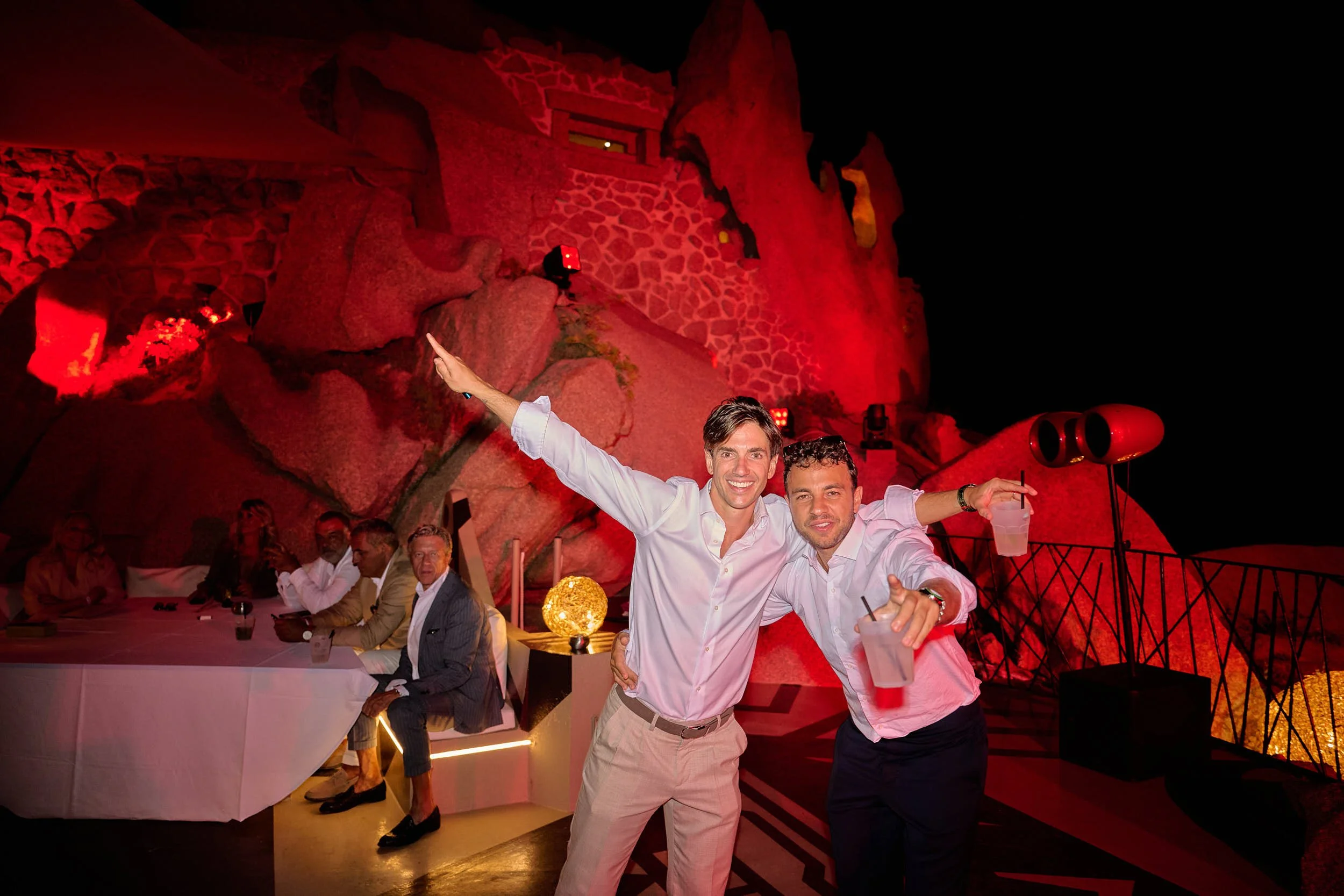 Groom and a friend pose with drinks as the party continues under dramatic red lighting