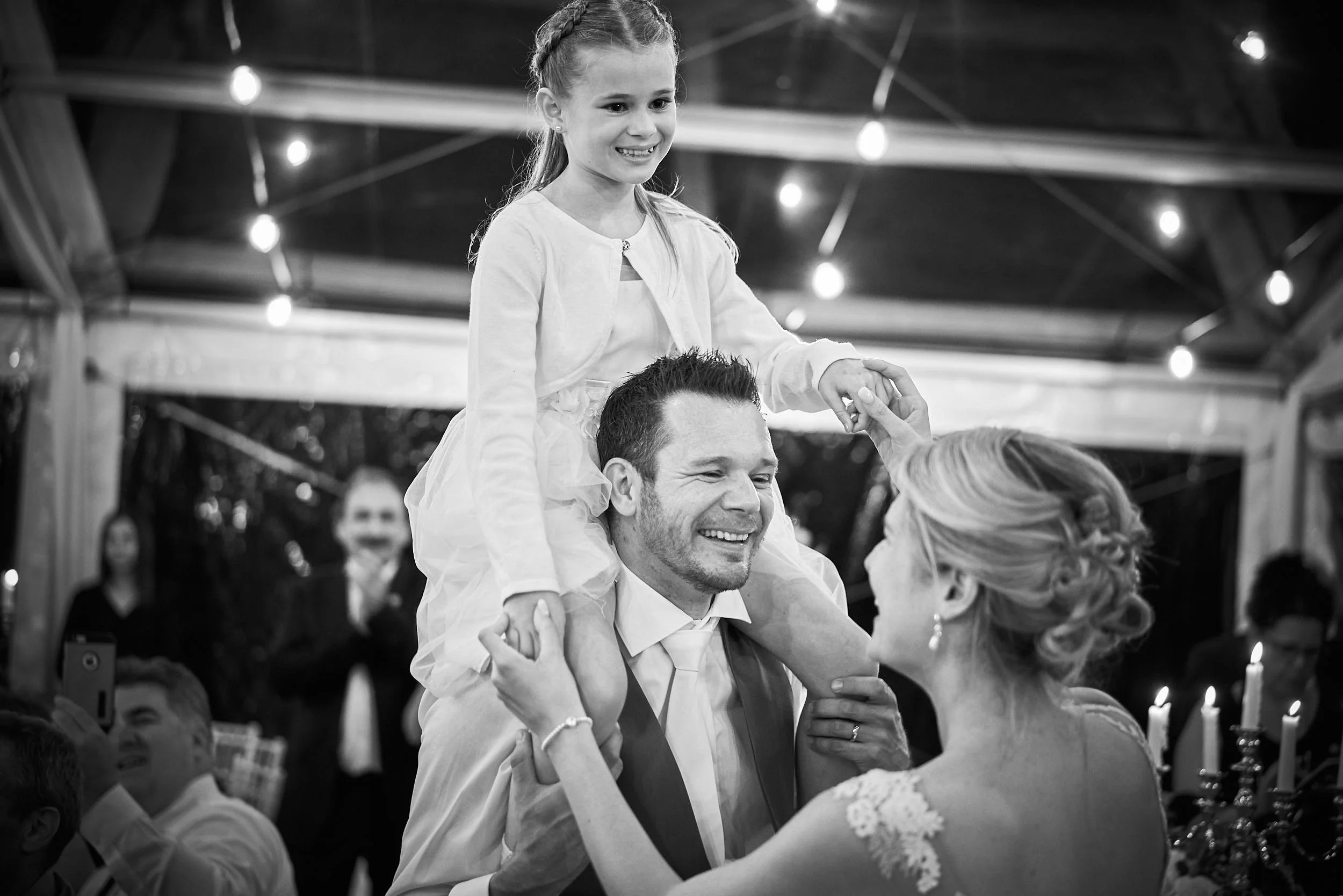 Black-and-white reception moment with a child on someone’s shoulders, smiling under bistro lights—family energy, candid and real.