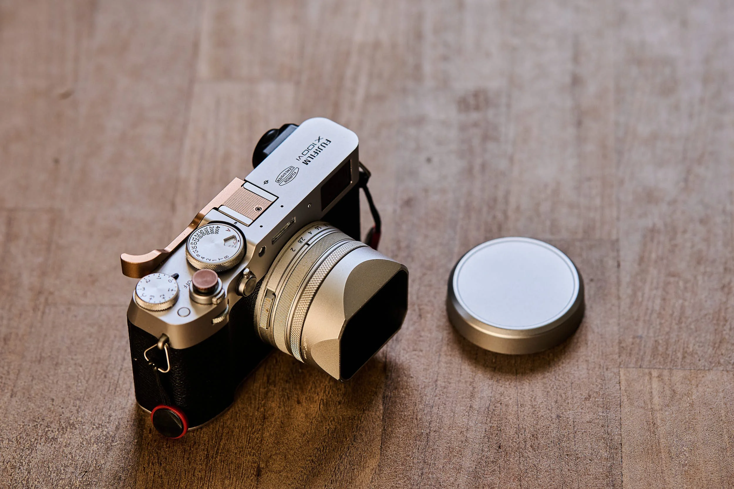 Fujifilm X100VI in silver with copper PolarPro thumb grip, soft-release button and square metal lens hood on a wooden surface, next to the front lens cap.