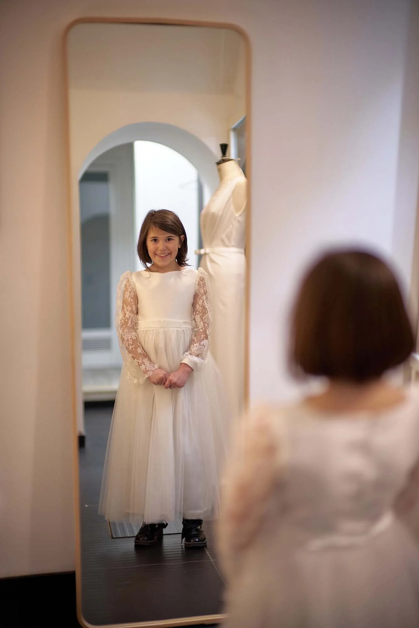 A young girl wearing a dress smiles at her reflection in a tall mirror, with a mannequin dress in the background.