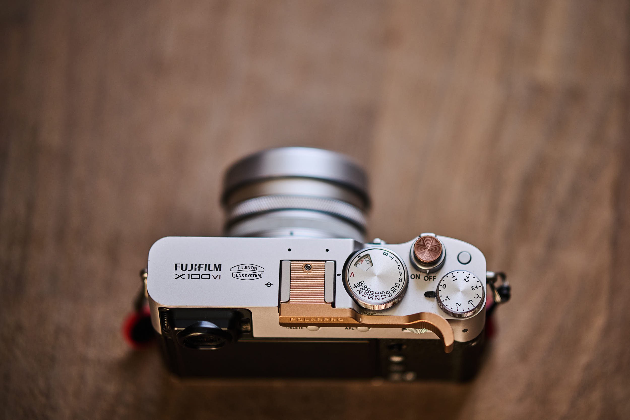 Top view of a silver Fujifilm X100VI with copper PolarPro thumb grip and soft-release button mounted on the shutter, resting on a wooden surface.