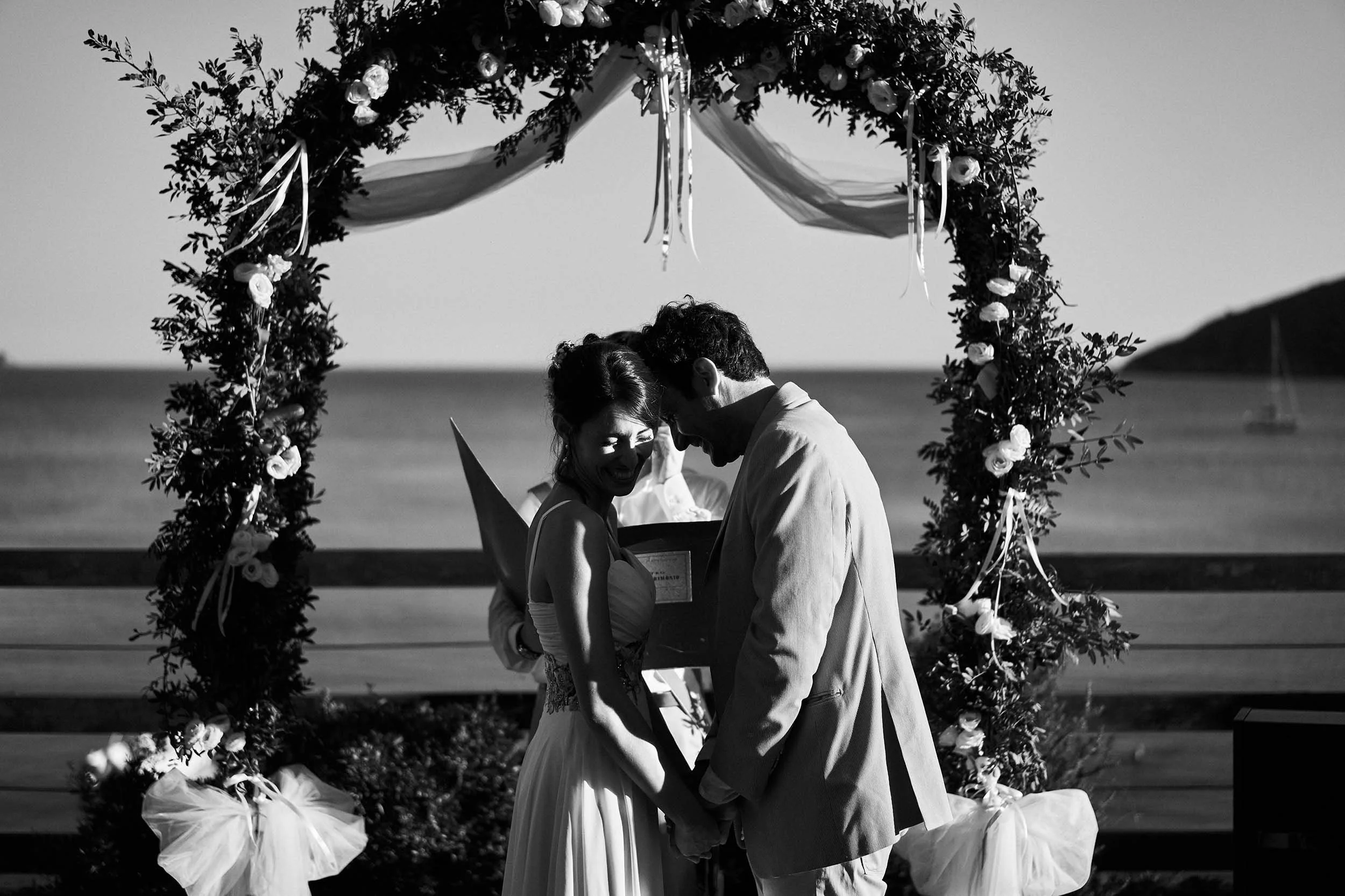 Black and white documentary wedding photography in Italy. A groom leans into his smiling bride under a floral arch by the sea, their intimate moment sheltered by deep, elegant shadows.