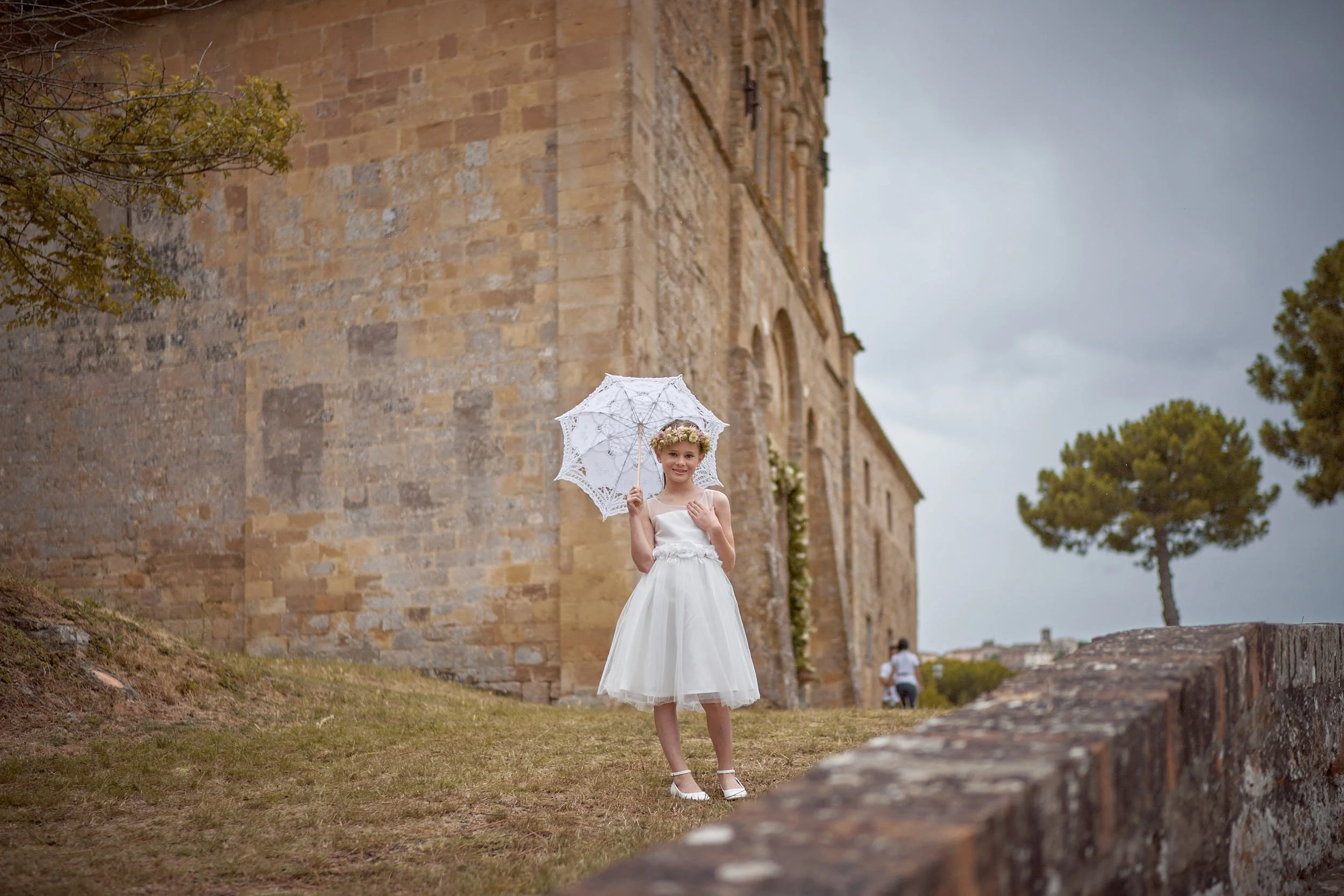 Flower girl wearing a flower crown holds a lace parasol in front of a Tuscan church.