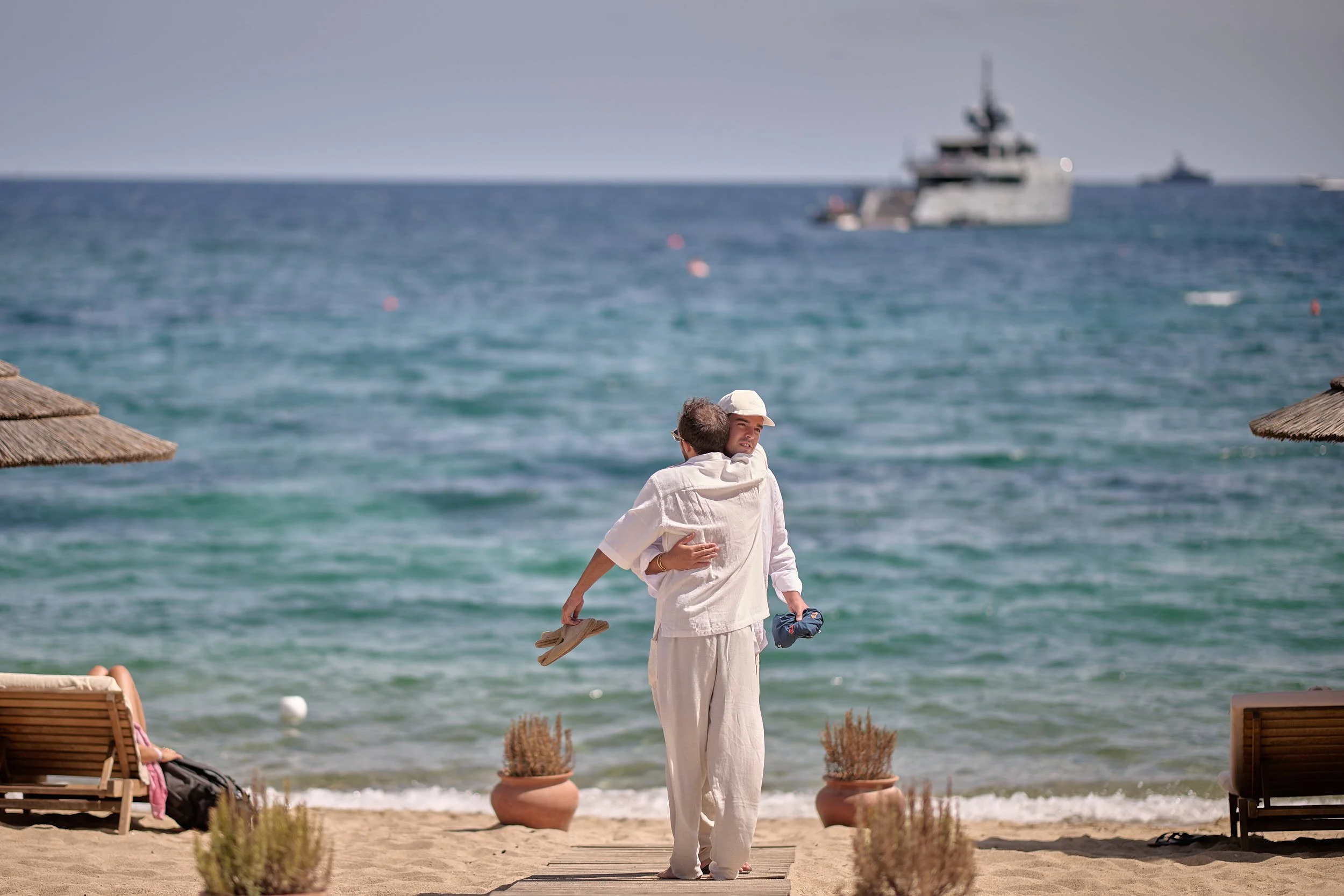 Two guests in white linen hug on a wooden walkway facing the sea, with beach umbrellas and a yacht offshore on the Costa Smeralda.