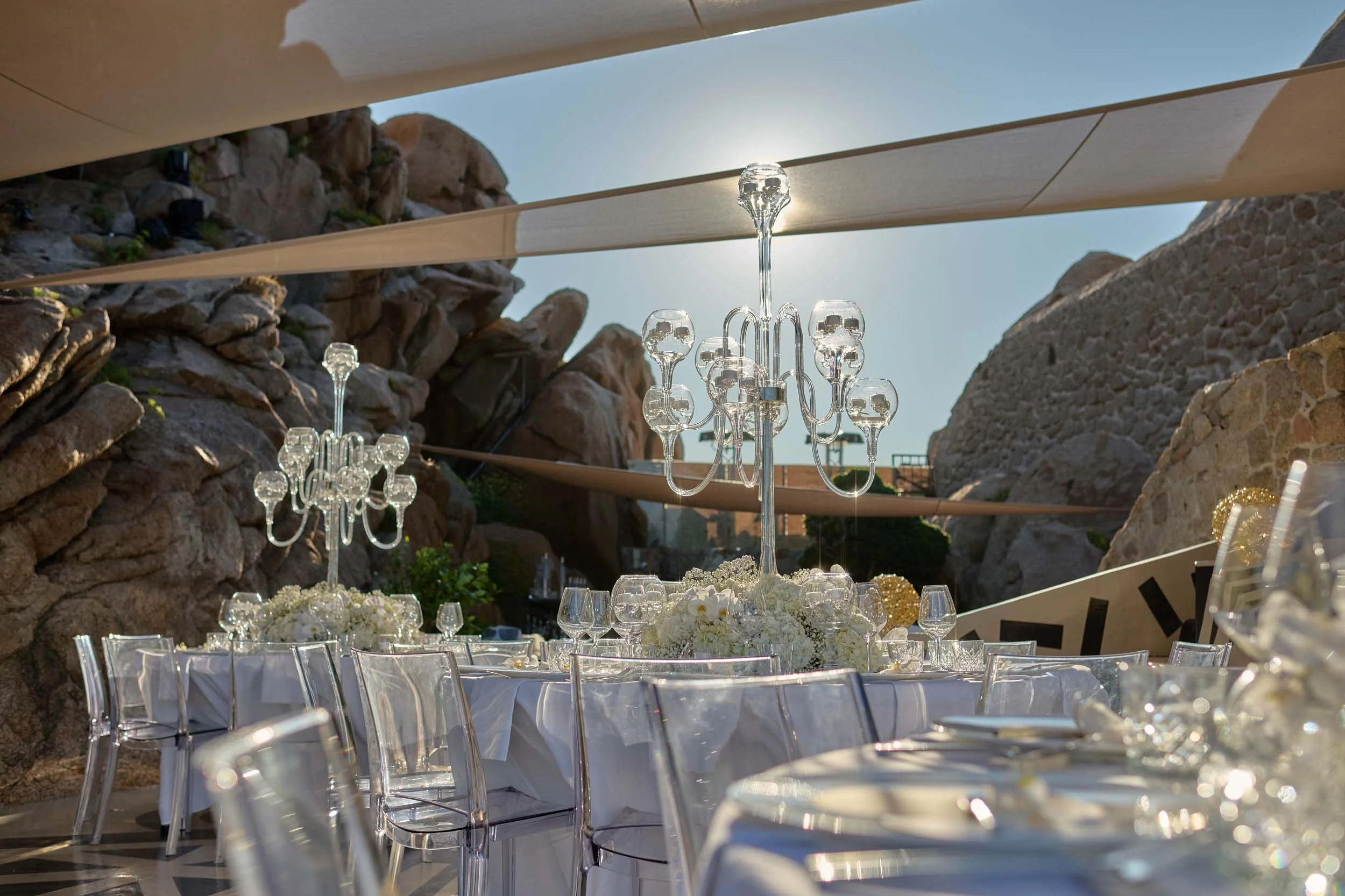 Reception tables with white linens, clear chairs and glass candelabras set among granite rocks at The Rock, Phi Beach, Costa Smeralda.