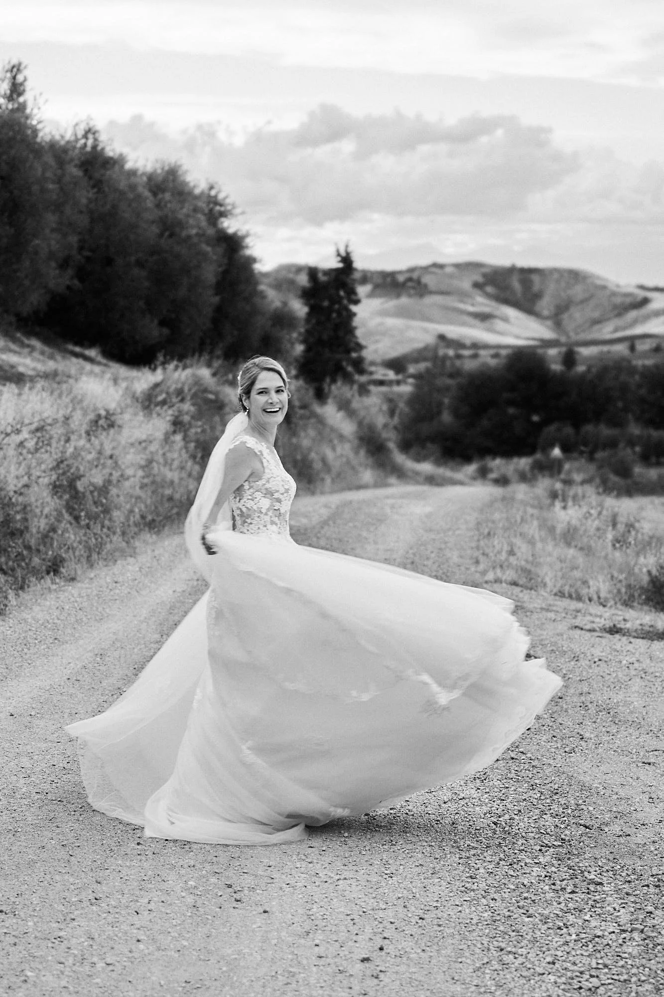 Black-and-white portrait of the bride spinning on a gravel road with Tuscan hills in the background, airy movement and cinematic space.