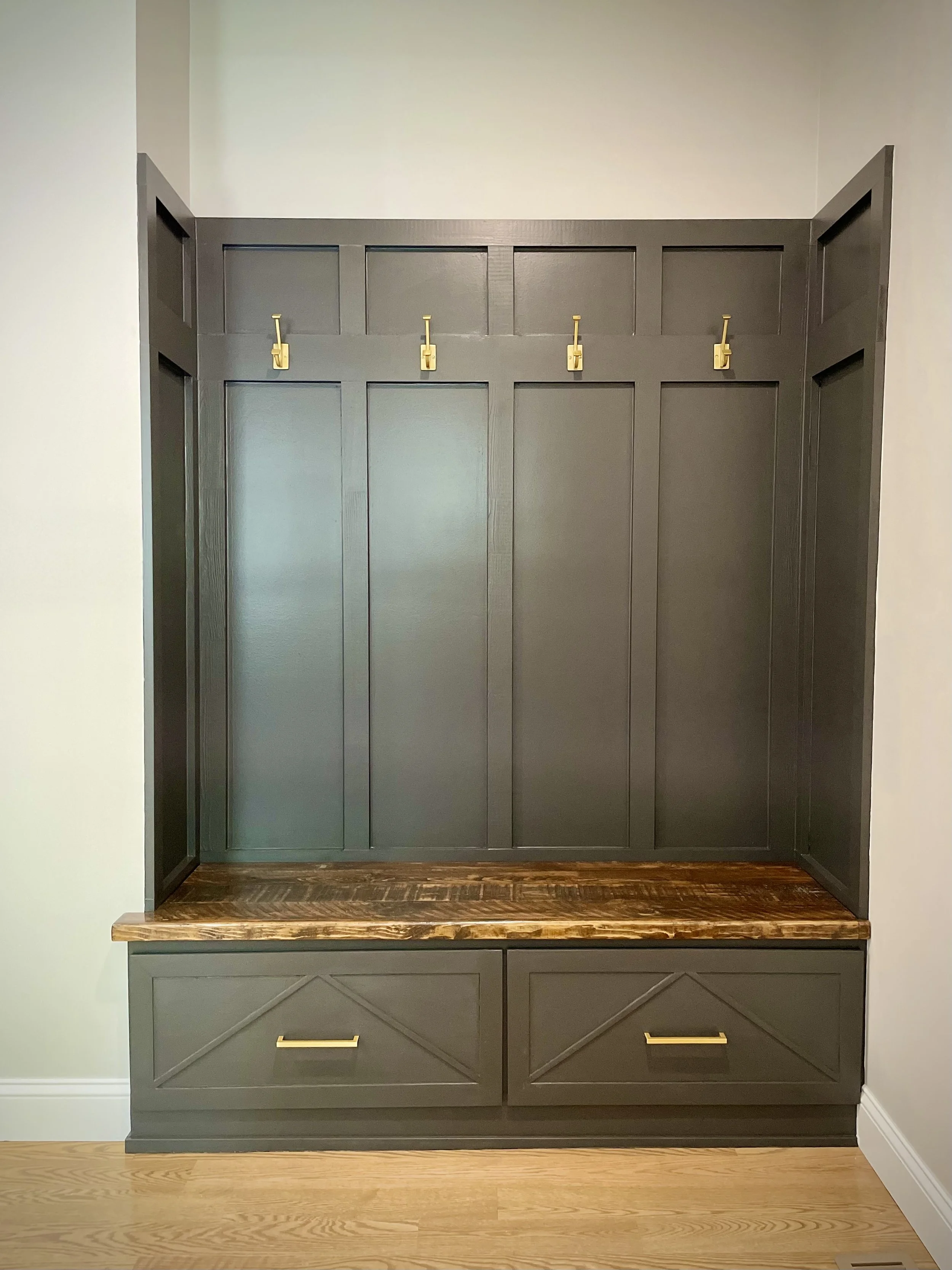 A gray wooden hall tree with a built-in bench and storage drawers. It has a back panel with four gold hooks for coats or bags. A wooden seat top and two large storage drawers with gold handles below.