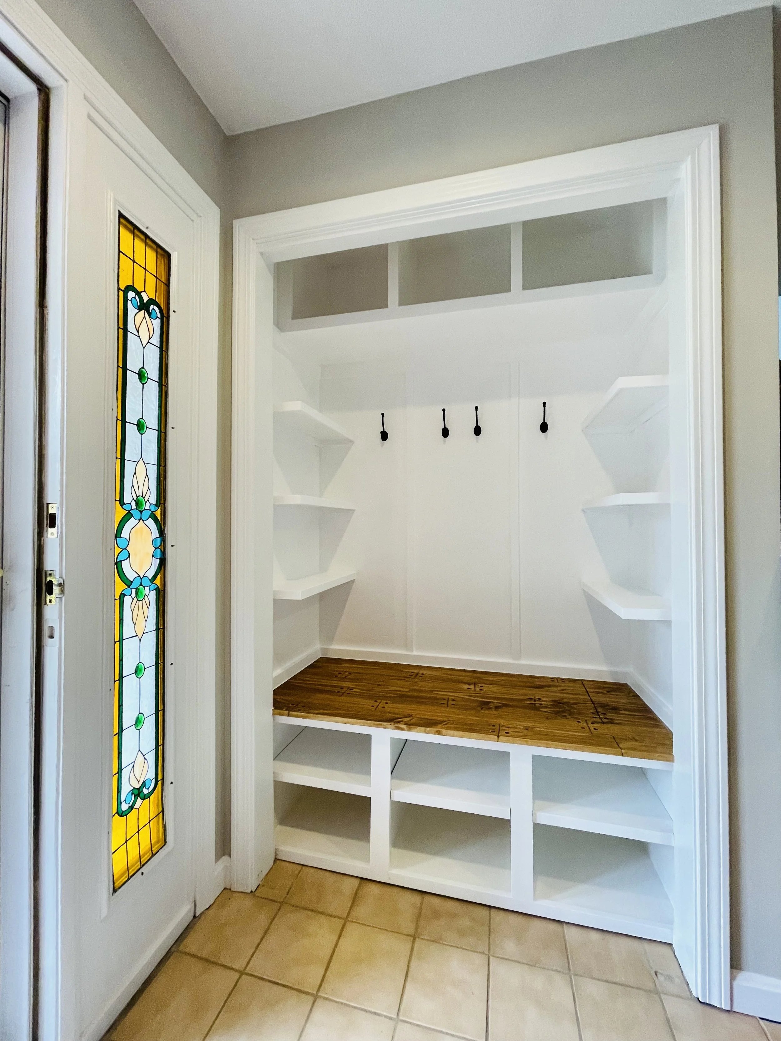 Empty mudroom with white built-in shelves, cubbies, black hooks, a upper window, and a wooden bench with cubbies underneath, next to a stained glass door with yellow, blue, green, and white designs.