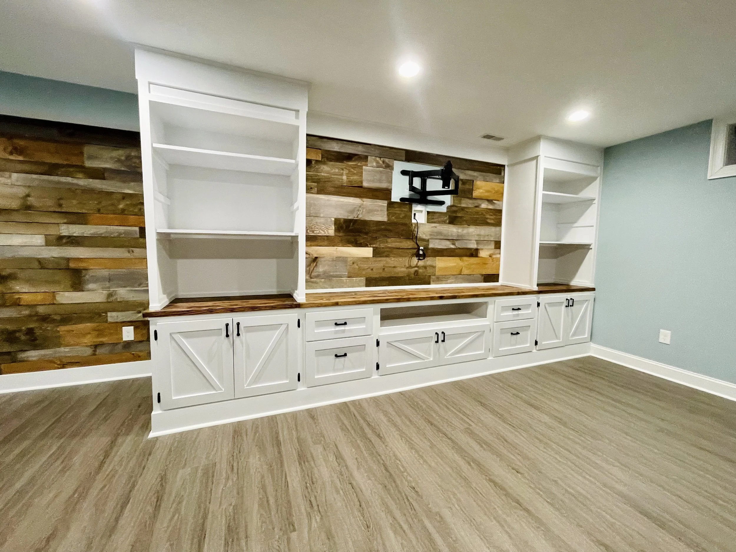 Empty built-in white cabinet with open shelves and drawers, a wooden countertop, and a wood-paneled accent wall in the background.