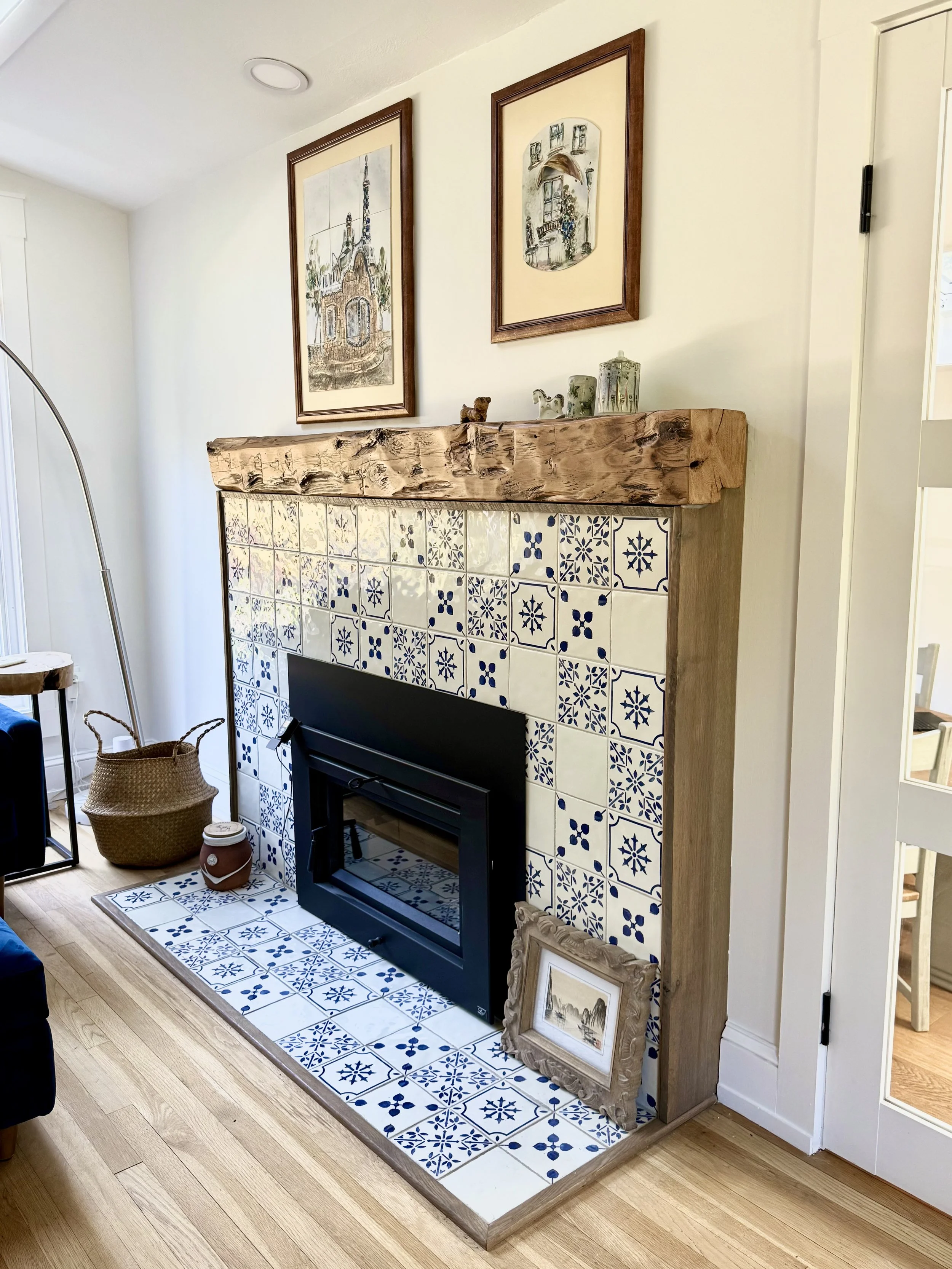 A cozy living room corner featuring a fireplace surrounded by blue and white decorative tiles with geometric patterns, topped with a rustic wooden mantle. Above the mantle are three framed artworks. To the left, there is a woven basket, and below it, a decorative jar. To the right of the fireplace, an ornate picture frame with landscape art is leaning against the wall.