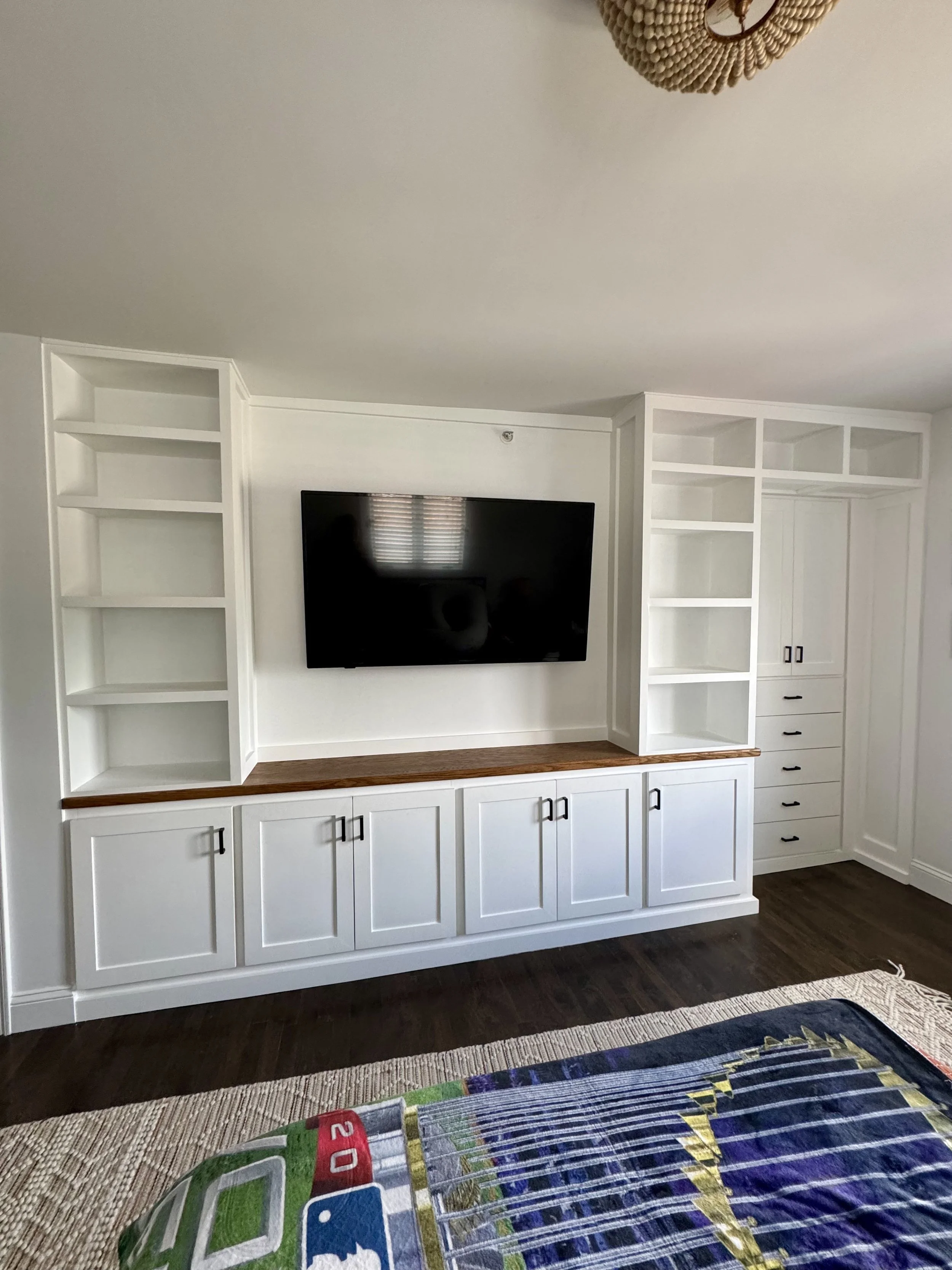 A living room with a white built-in entertainment center, a large flat-screen TV, and dark hardwood floors. There is a colorful bedspread in the foreground.