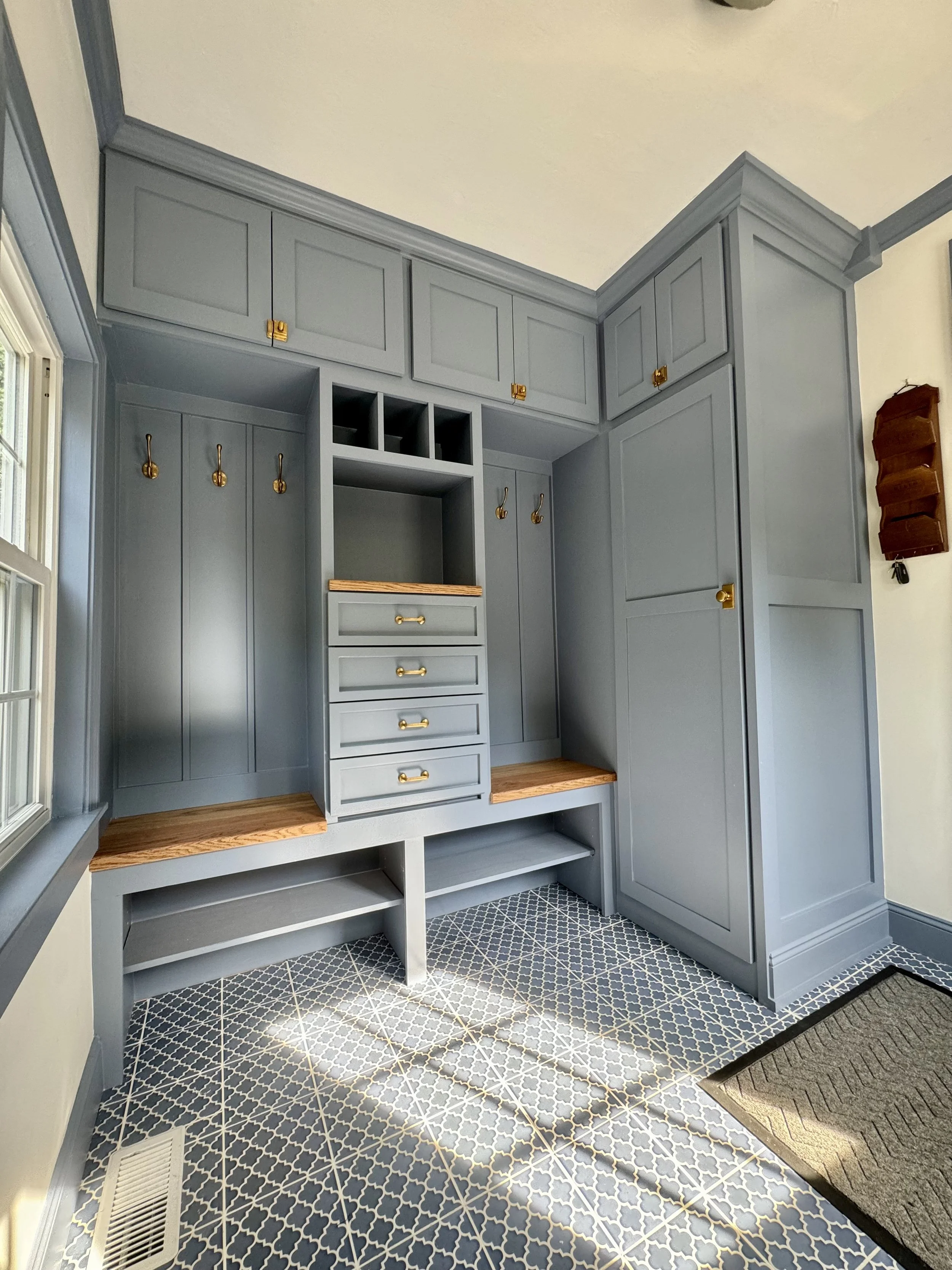 A built-in light blue storage and seating unit with cabinets, drawers, hooks, and wooden benches in a corner of a room with patterned floor tiles and a window.
