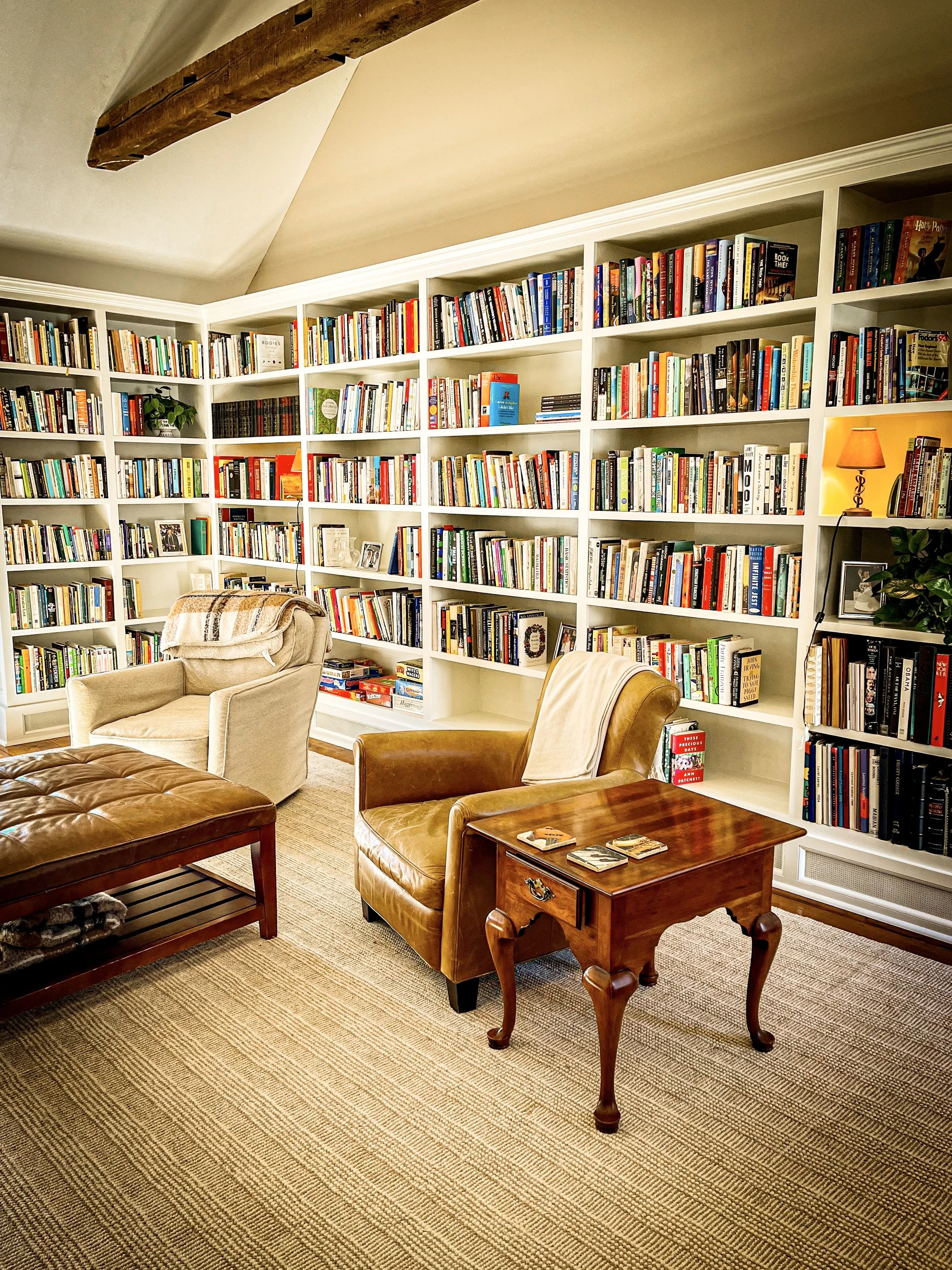 Living room with large white bookshelves filled with colorful books, two armchairs, a table with a few cards, a padded ottoman, and a beige carpet. A lamp and framed photo are on the bookshelf.
