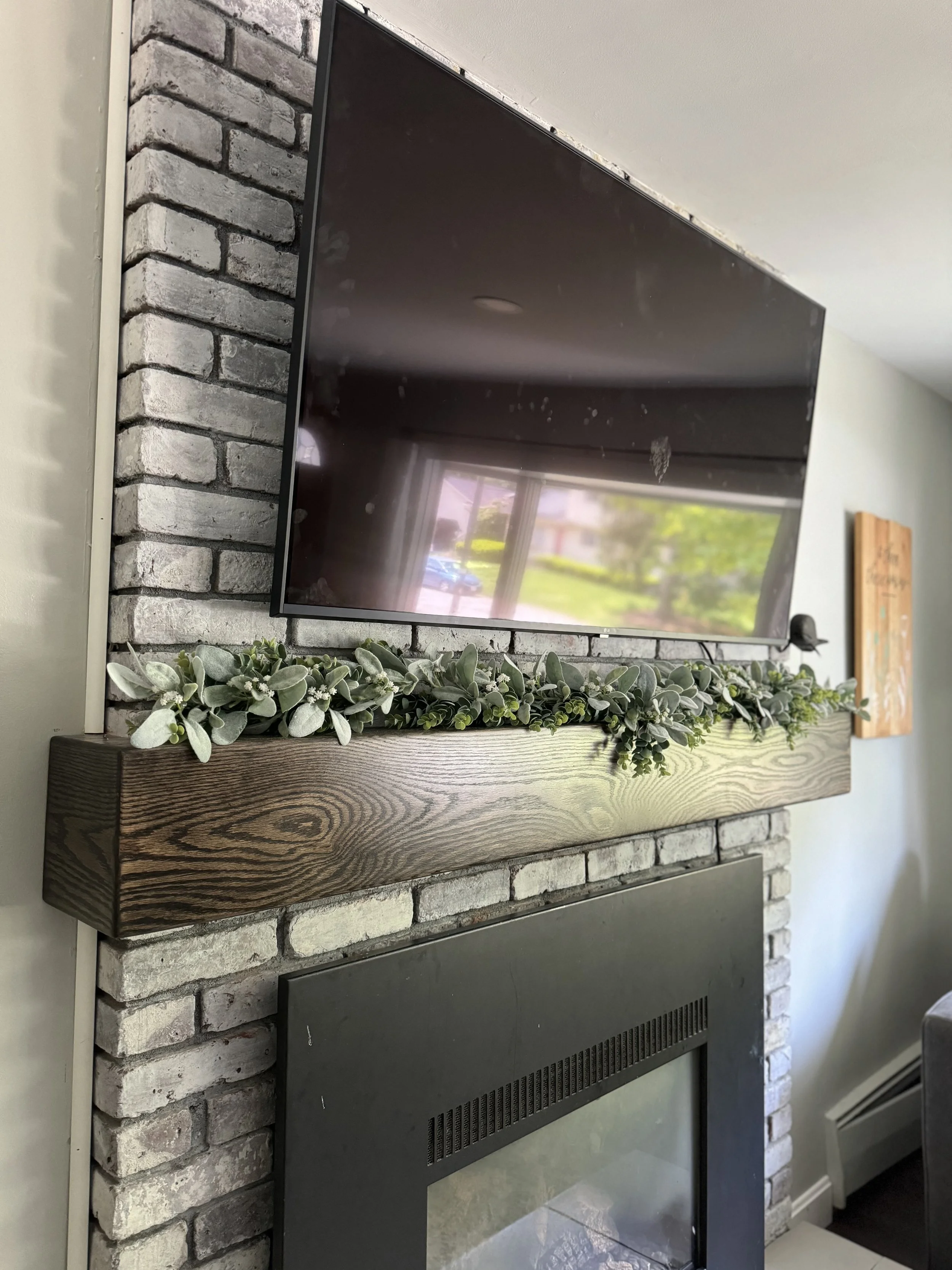 A wall-mounted flat-screen television above a brick fireplace with a wooden mantel and a decorative garland of greenery. There is a window reflecting outside scenery in the TV screen.