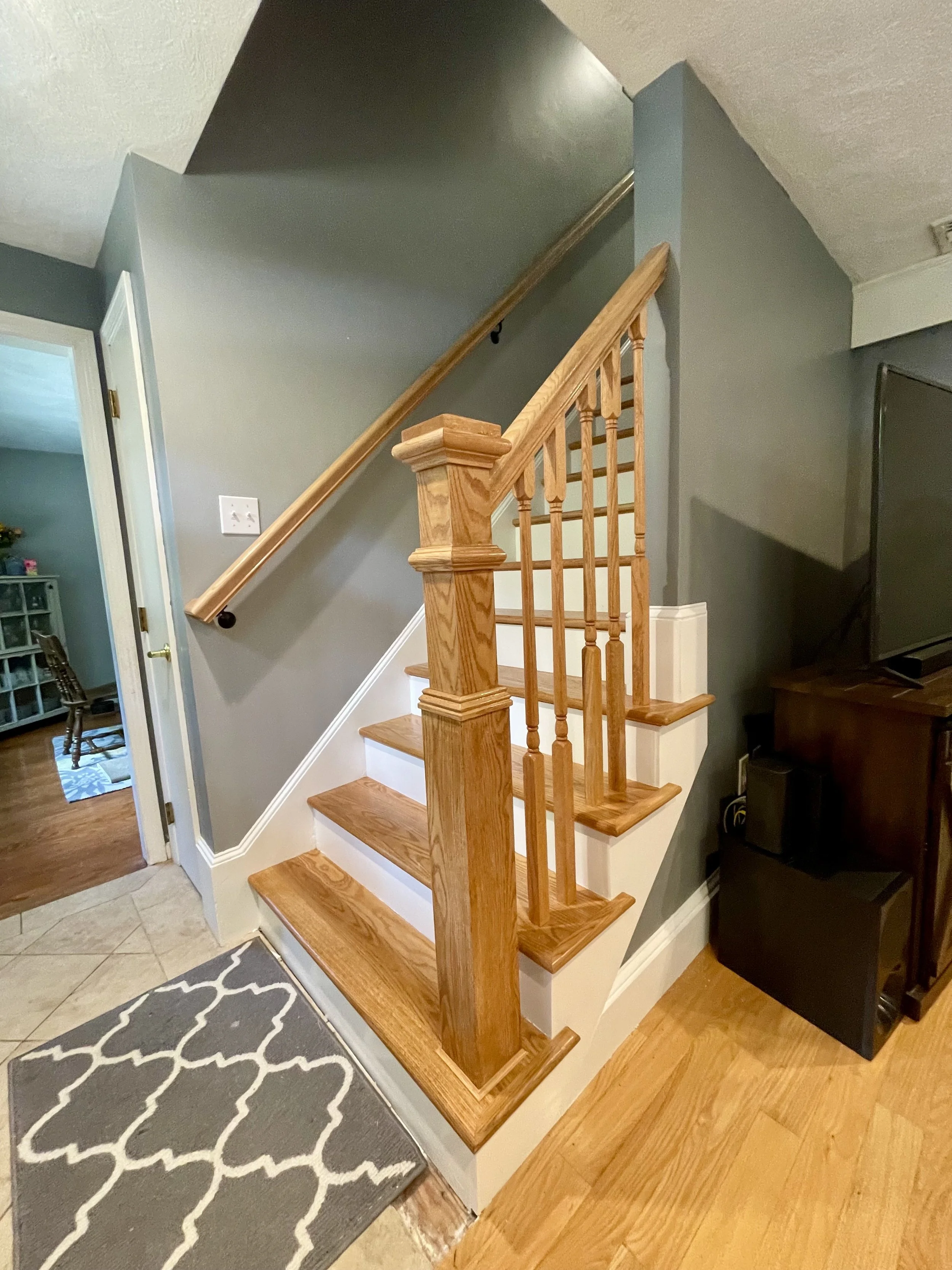 A wooden staircase with a curved handrail and balusters near a corner in a living room, with a decorative rug on the hardwood floor and a nearby television and speaker.