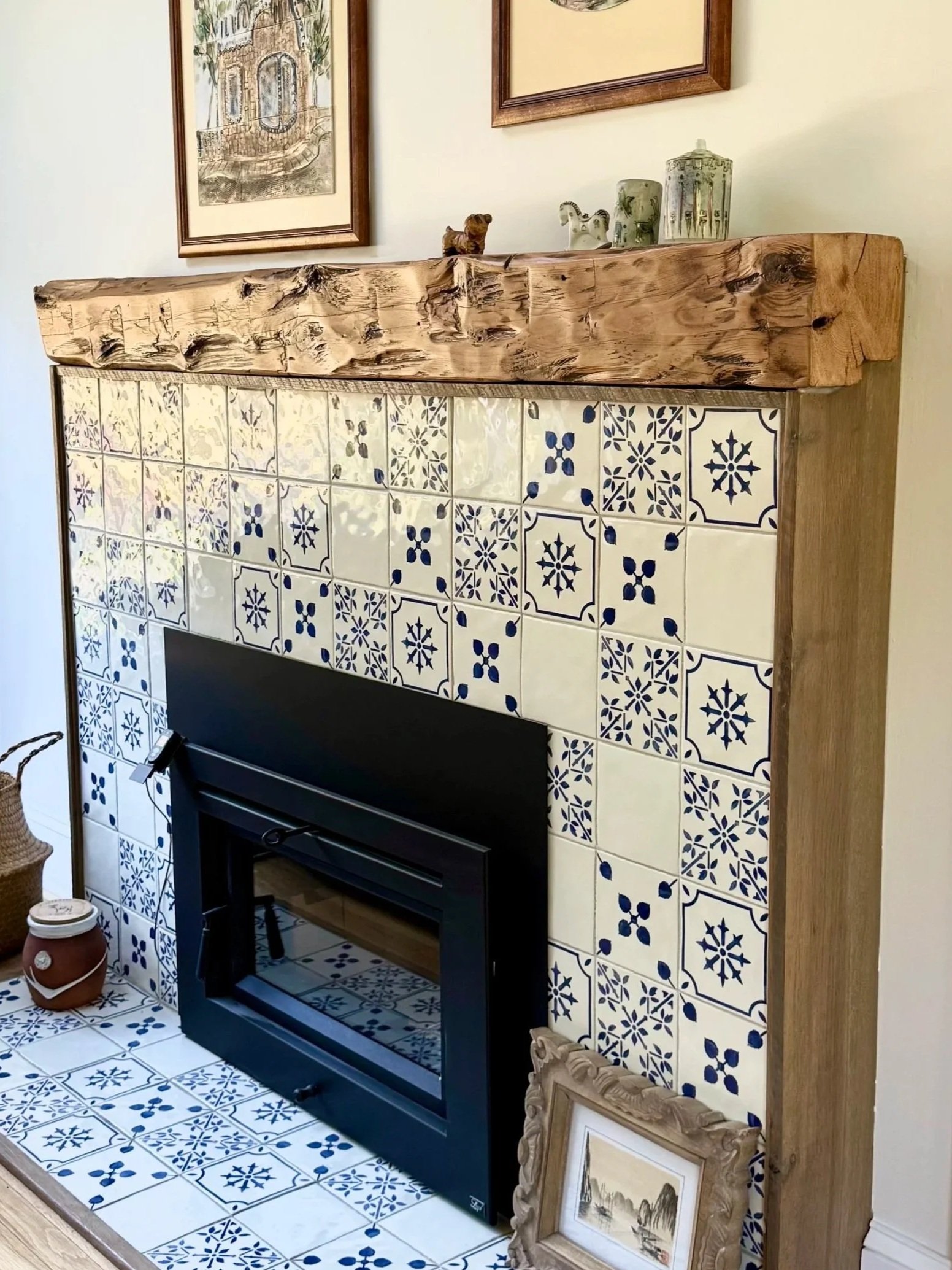 Decorative fireplace with a wooden mantle, tiled surround with blue and white patterns, framed artwork above, and a small framed picture leaning against the side.