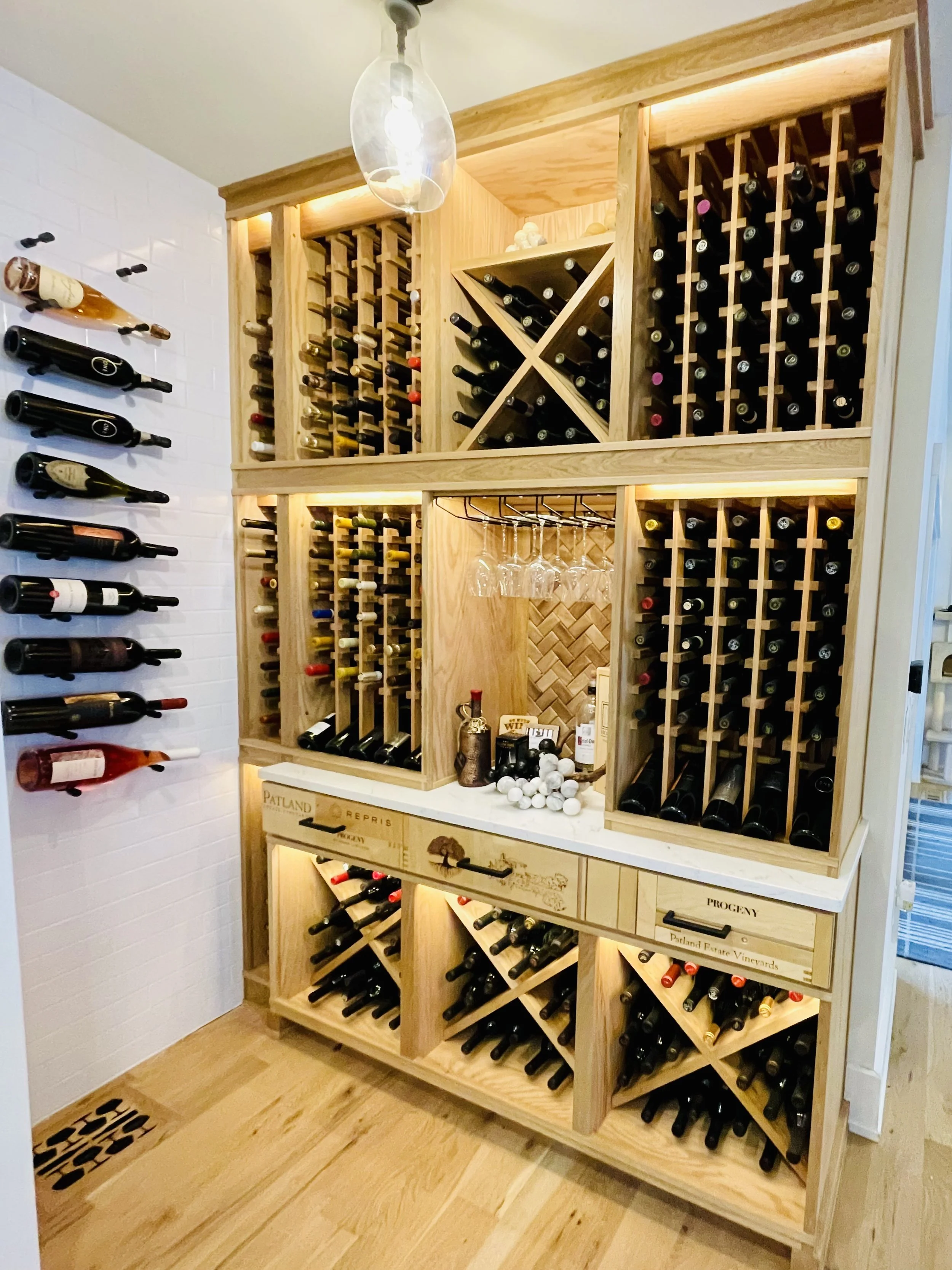 A wooden wine cellar with multiple shelves filled with bottles of wine, includes a countertop with wine accessories and glasses hanging.