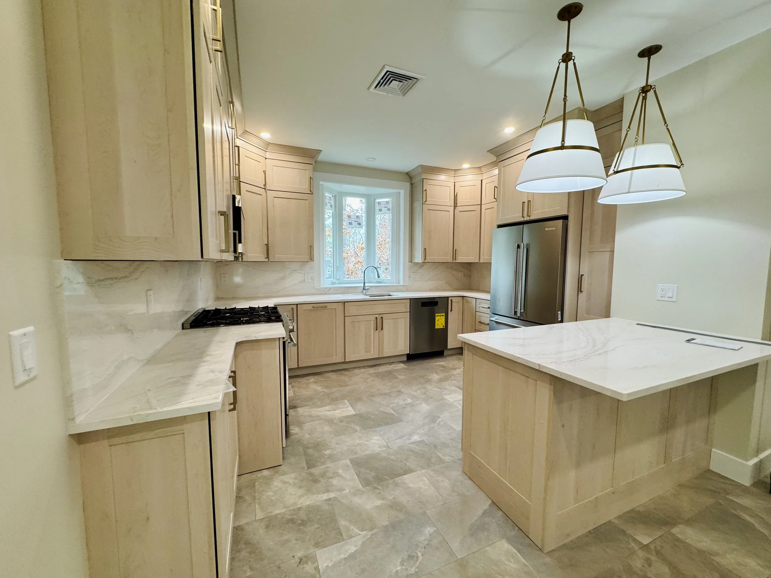 Kitchen with light wood cabinets, marble countertops, stainless steel refrigerator, gas stove, and pendant lighting over a central island.