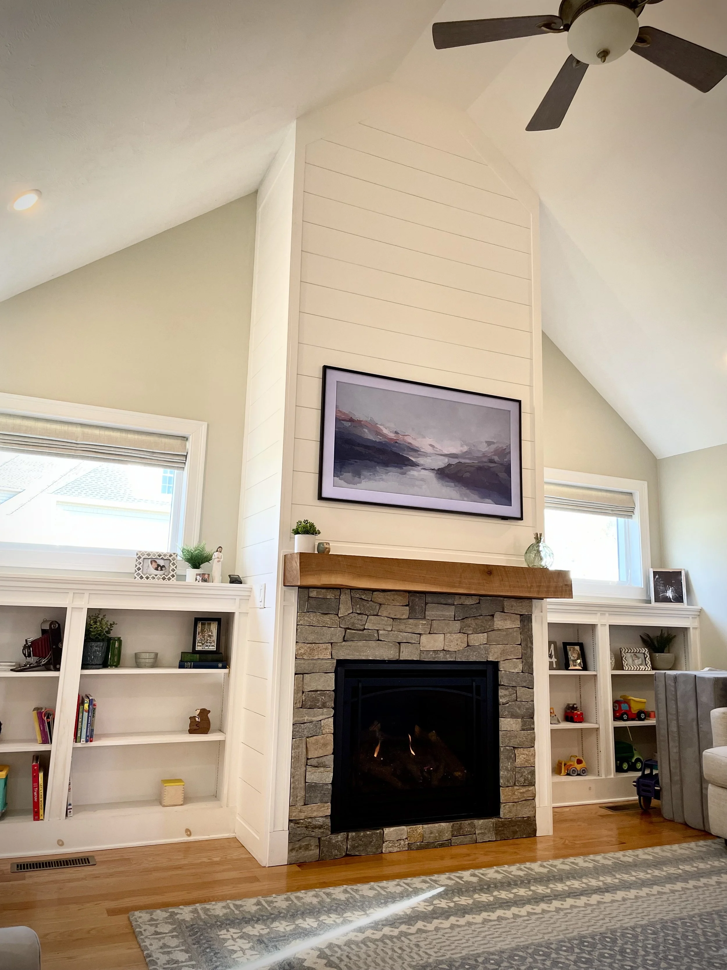 Living room with a stone fireplace, mounted landscape painting above, white built-in shelves with books and decor, windows on each side, hardwood flooring, patterned area rug, ceiling fan.