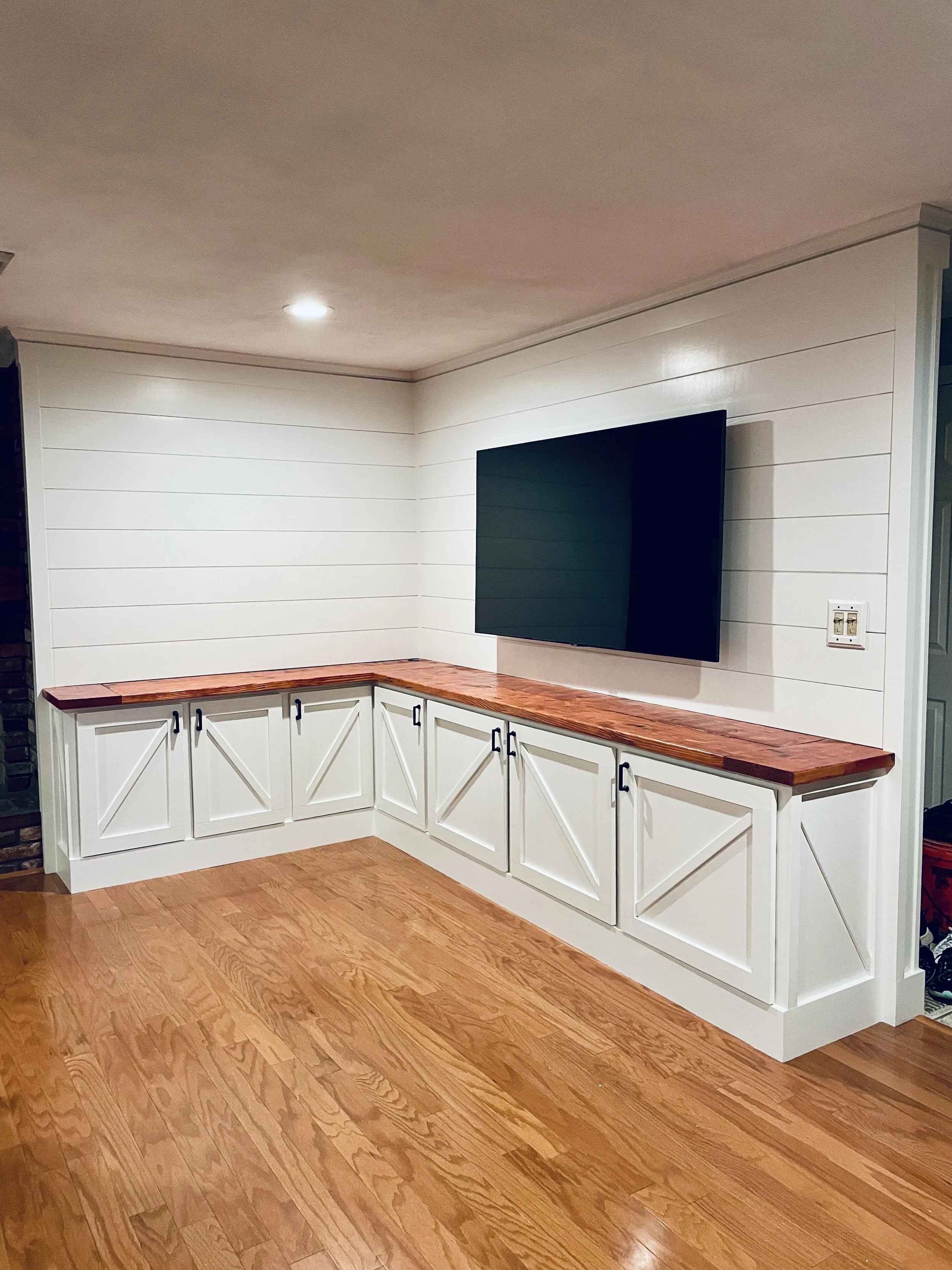 Living room corner with a built-in white cabinet and a mounted flat-screen TV on white paneled wall, hardwood floor.