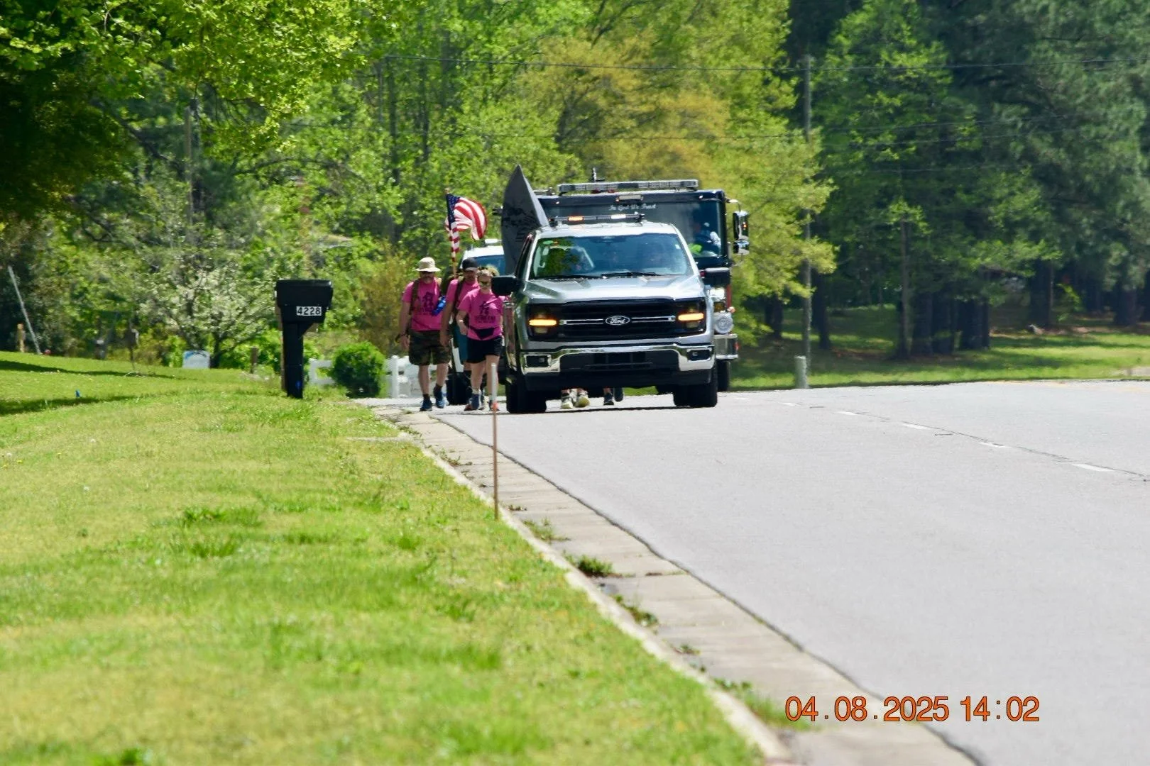 Group of people walking on the sidewalk next to parked police vehicle, with trees and grassy area in the background, date and time stamp at the bottom.