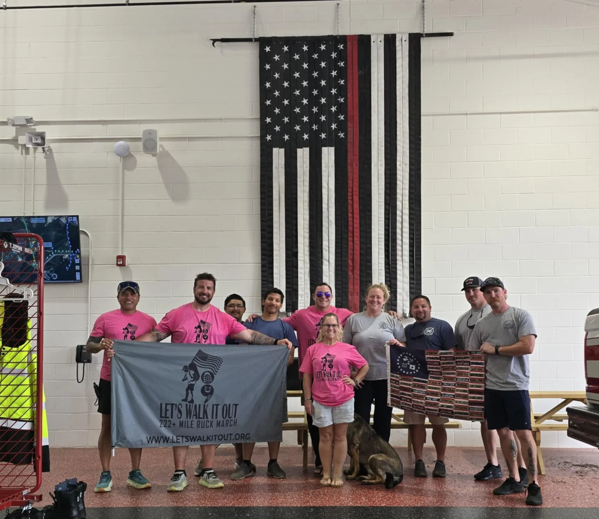 Group of people standing together indoors, holding flags for a march, with a large American flag-themed wall hanging behind them. Some individuals are wearing pink shirts, and there is a dog sitting in front. A TV screen and equipment are visible to the left.